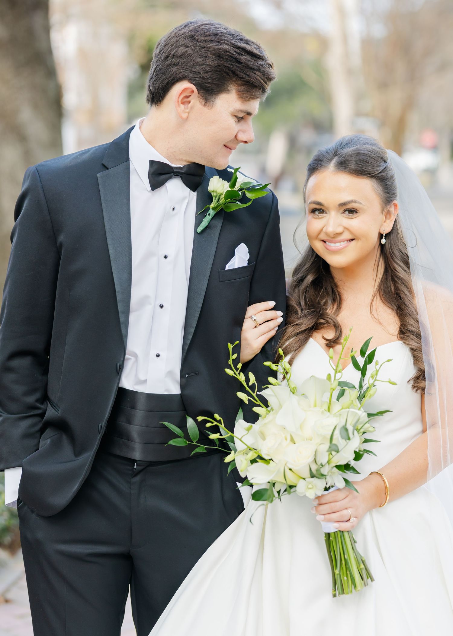 Elegant bride and groom pose together in formal attire with white floral bouquet in outdoor setting.