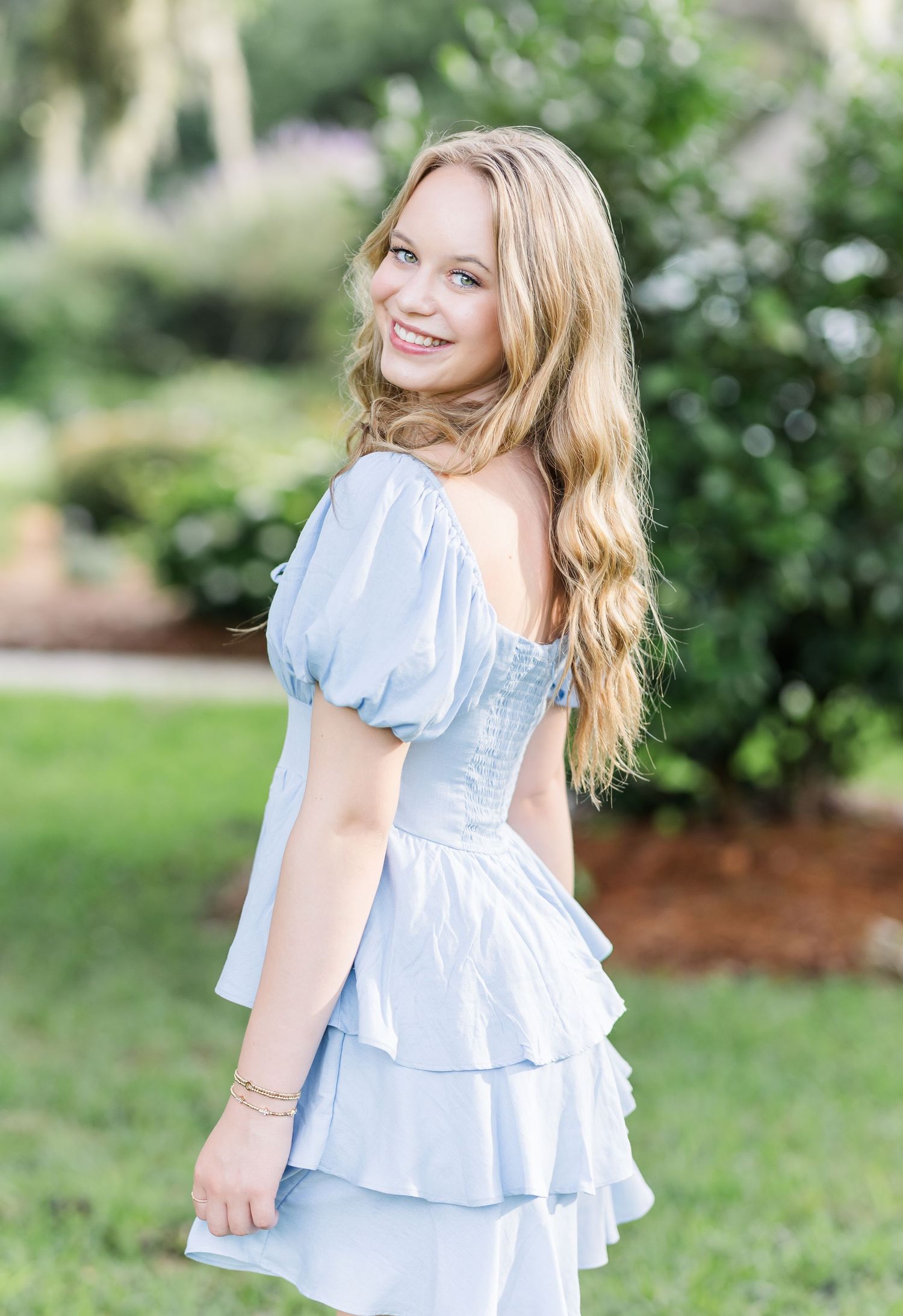 Young child wearing a light blue ruffled dress spins and plays in an outdoor garden setting on a sunny day.