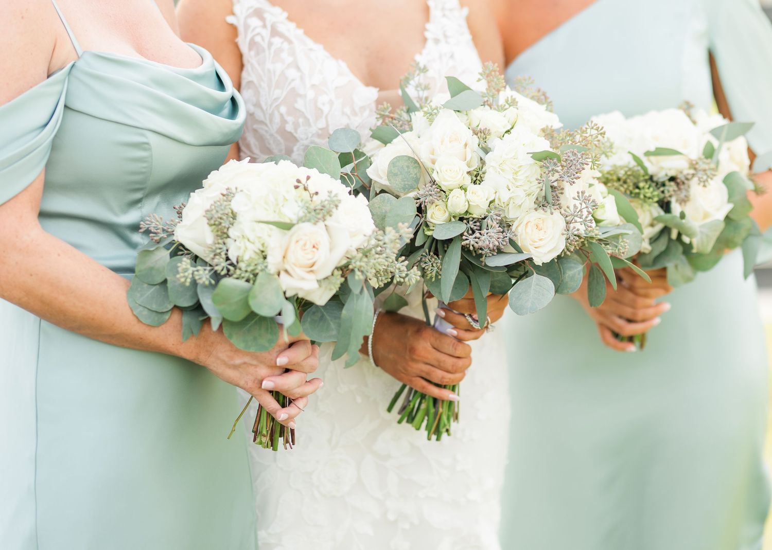 Wedding party members in mint green dresses hold elegant white and green floral bouquets during outdoor ceremony.
