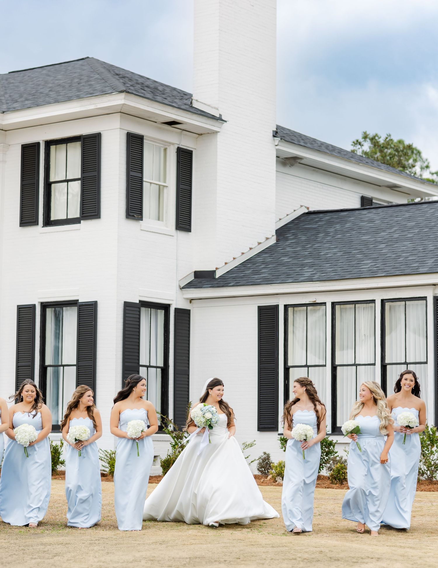 Bridesmaids in light blue dresses walking with bride in white ball gown outside white venue.