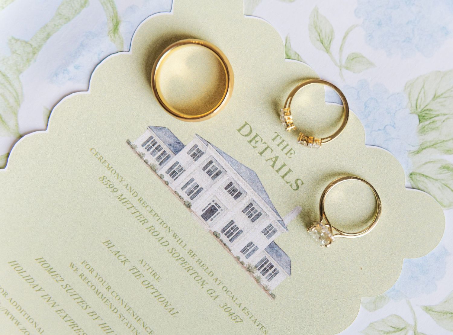 Gold wedding rings resting on an elegant wedding details card with a watercolor building illustration on floral background.