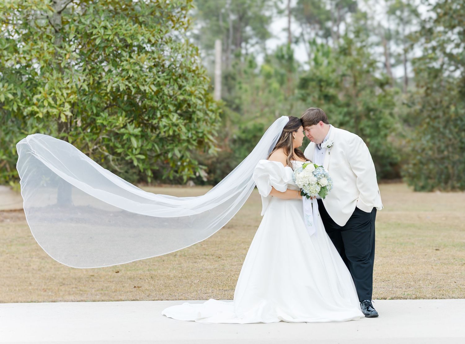 Bride and groom kissing with long veil flowing in the wind in an outdoor setting.