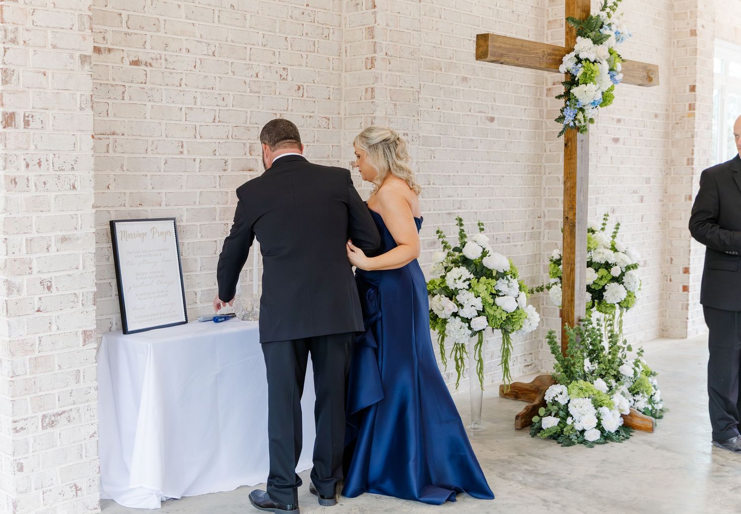 Couple in navy formal wear signing wedding registry near floral cross arrangement in white brick chapel.