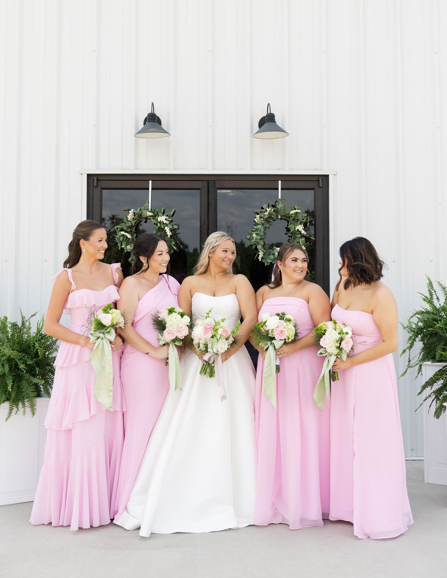 Bride in white gown with four bridesmaids in pink dresses holding bouquets outside a white barn venue.
