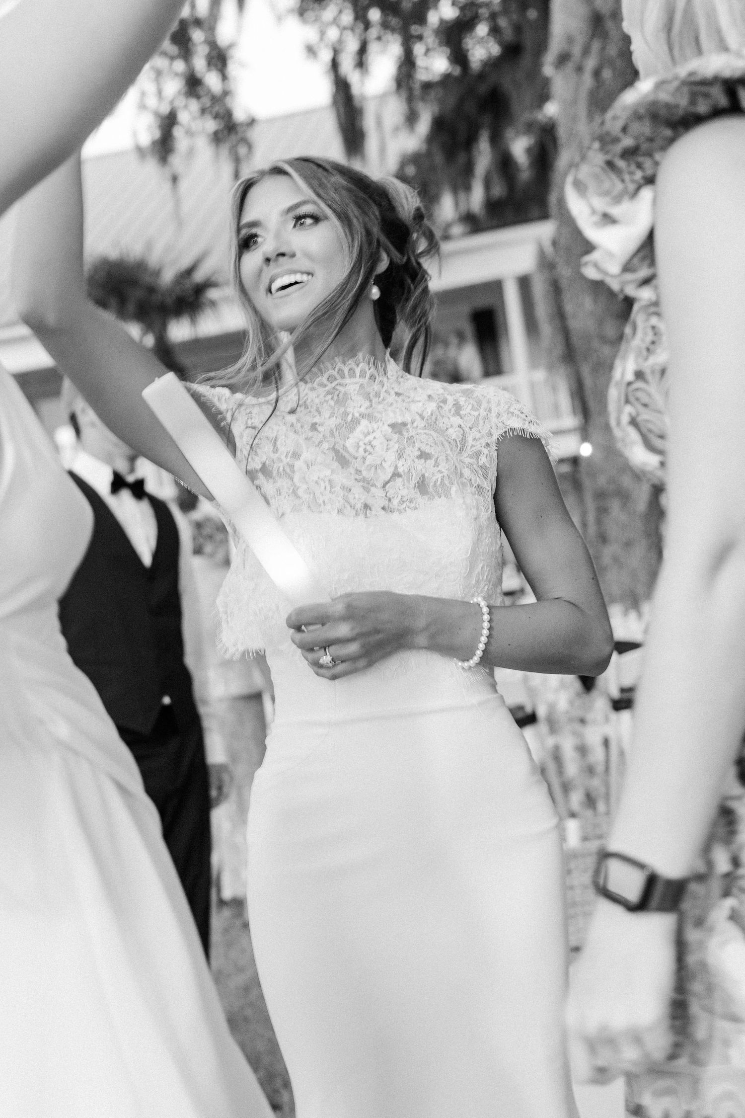 Black and white photo of smiling bride in lace gown holding glow stick during outdoor wedding reception.