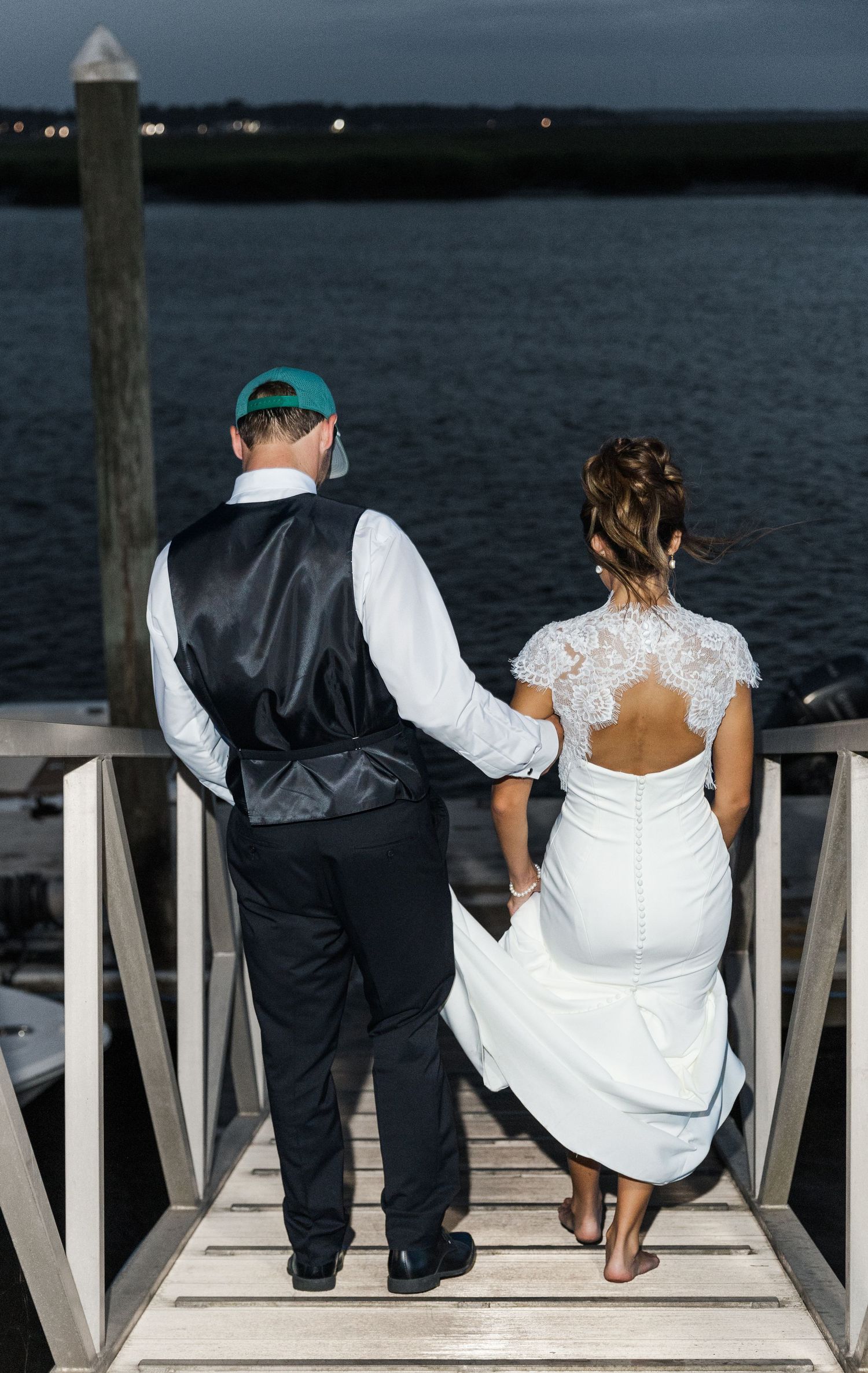 Bride and groom holding hands on a dock at dusk, overlooking calm water, bride in lace backless gown.