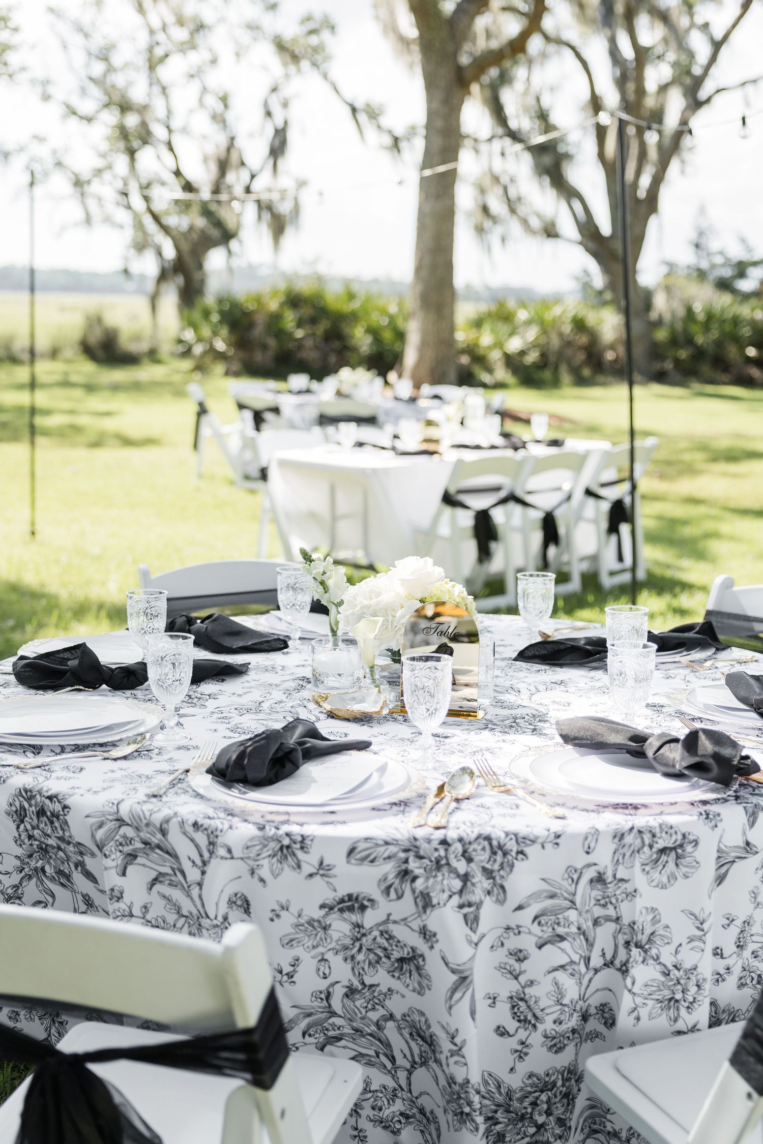 Elegant outdoor wedding reception with black and white floral tablecloths, black napkins, and white floral centerpieces on round tables.
