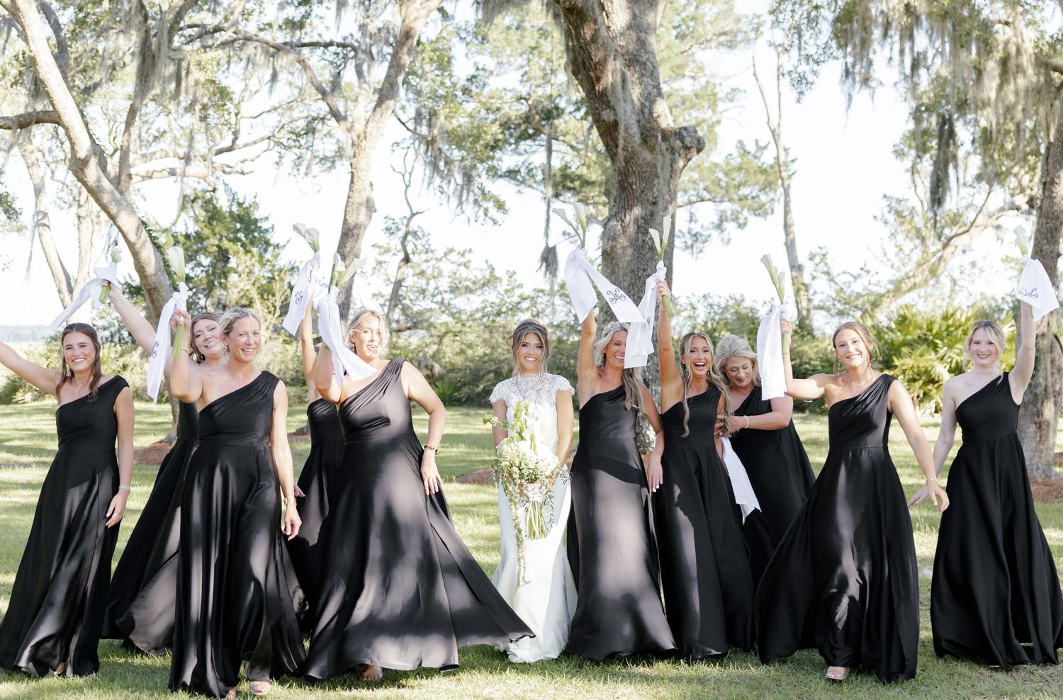 Bride in white gown with bridesmaids in black dresses raising white gloves outdoors under trees.