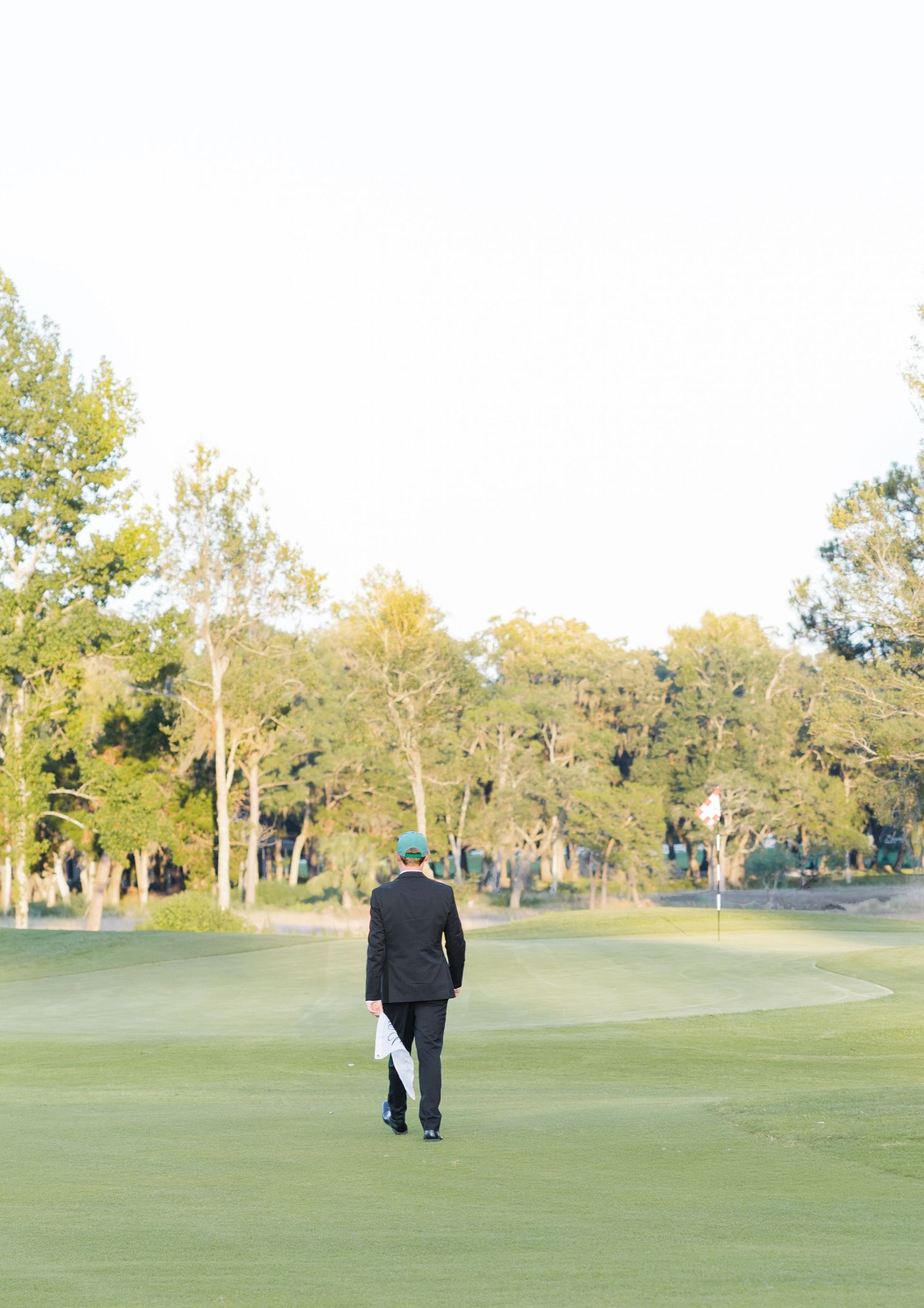 A golfer in a black suit and green cap walks across a lush golf course fairway surrounded by tall trees.