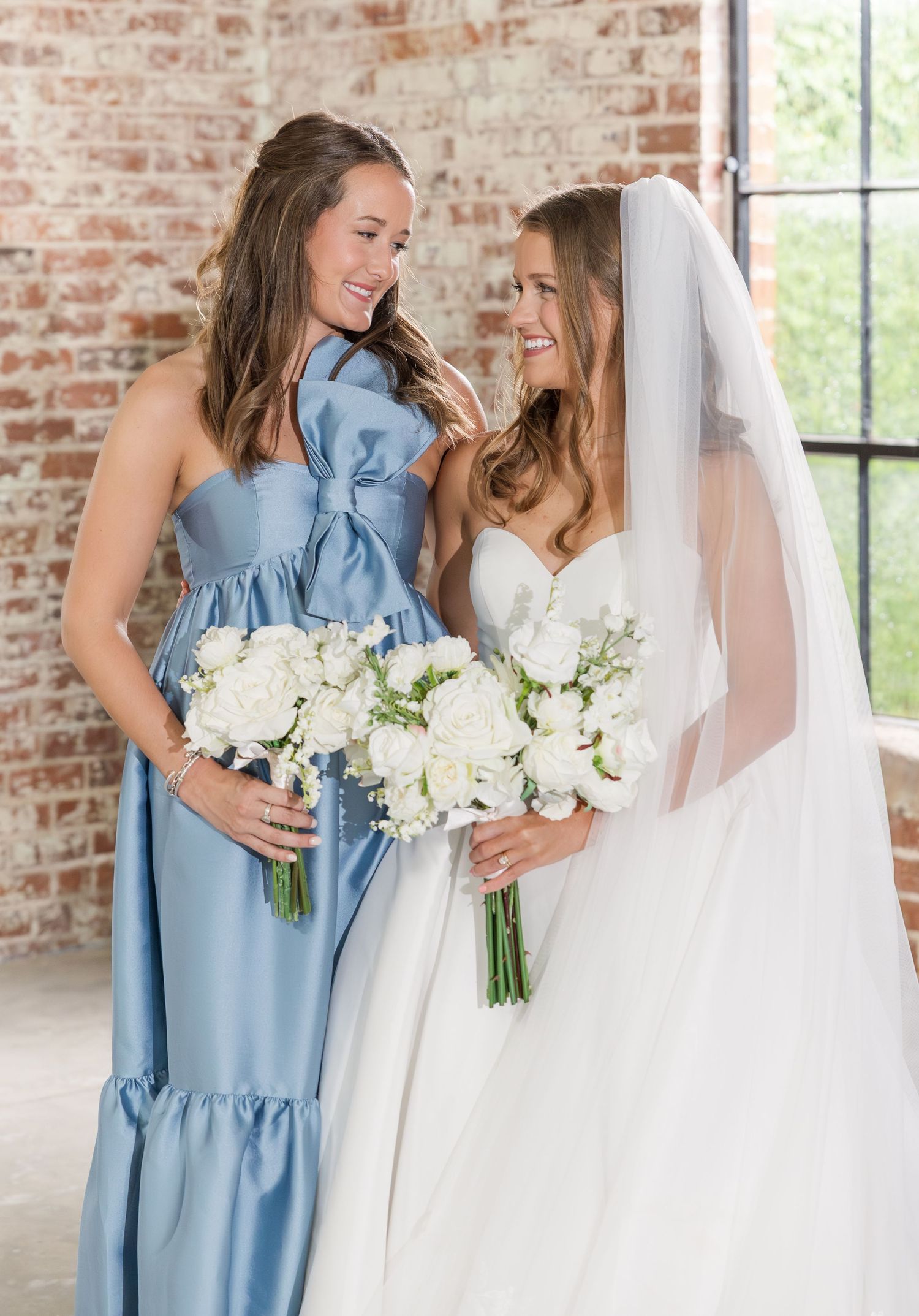 Bride in white gown and veil holds bouquet beside bridesmaid in blue dress, both smiling in rustic brick venue.