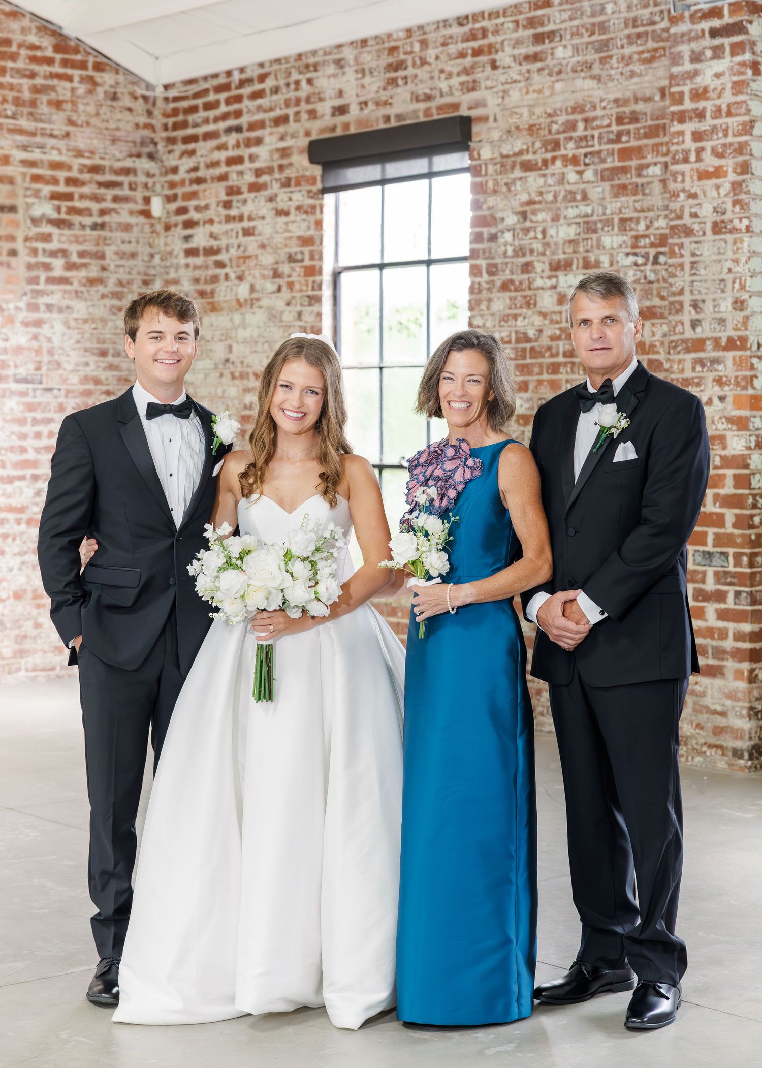 Wedding family portrait with bride, groom, and parents against exposed brick wall backdrop.