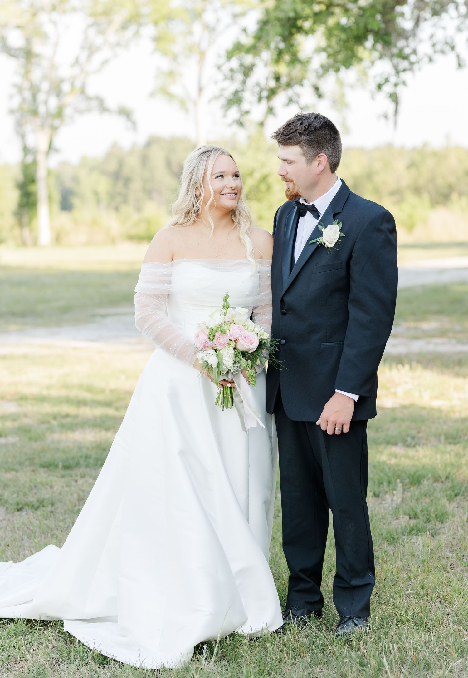 Smiling bride in elegant white A-line gown with groom in black tuxedo on green lawn.