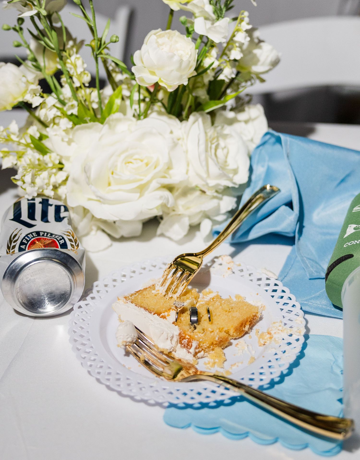 Wedding reception dessert plate with cake slice, gold fork, blue napkin, white roses, and Miller Lite can.