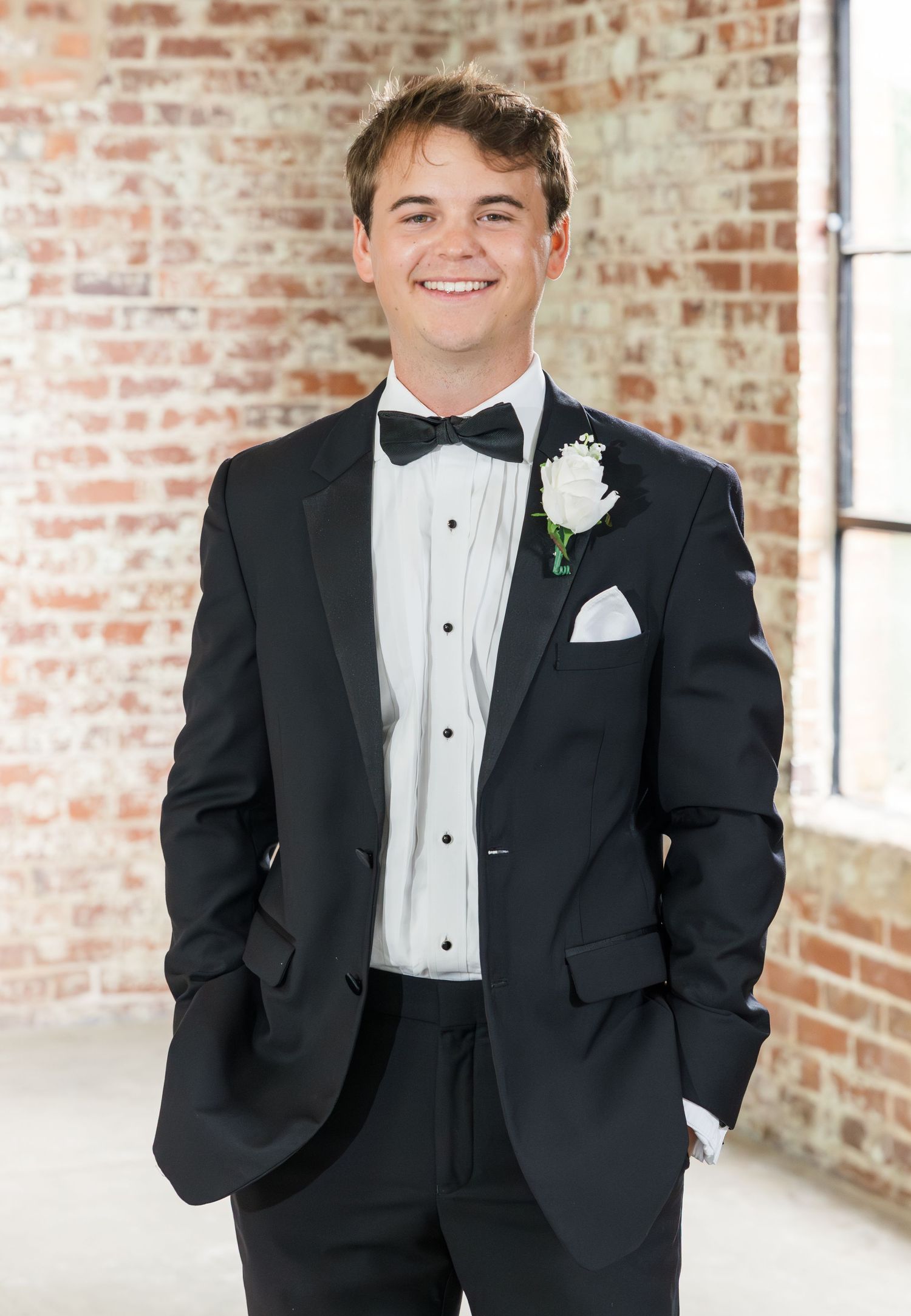 Young groom smiling in black tuxedo with bow tie and white boutonniere against brick wall.