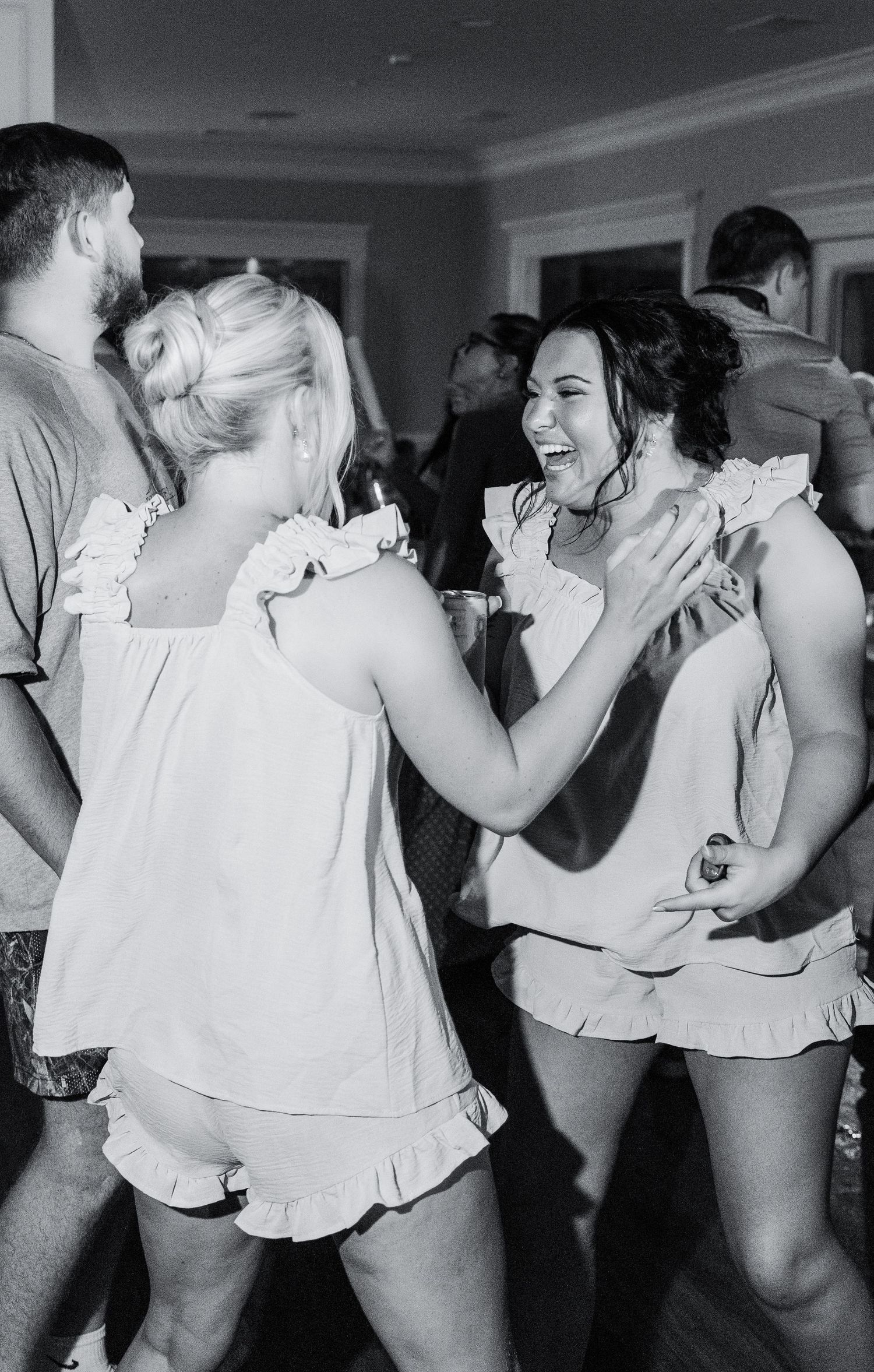 Two women in matching white ruffled outfits laugh and dance together at a lively wedding reception party.