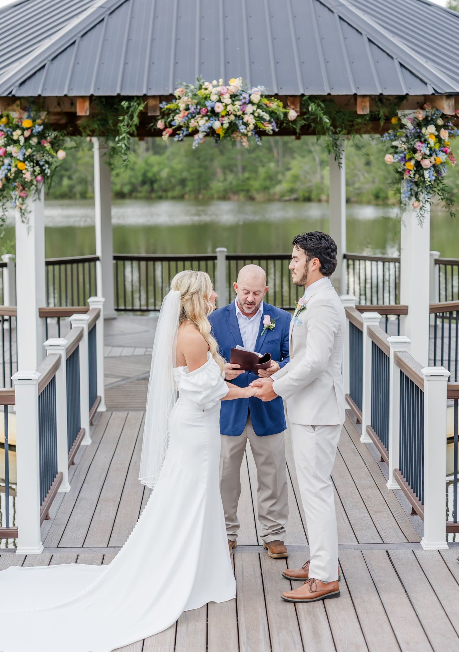 Bride and groom exchange vows at a gazebo ceremony overlooking a lake with floral arch decorations.