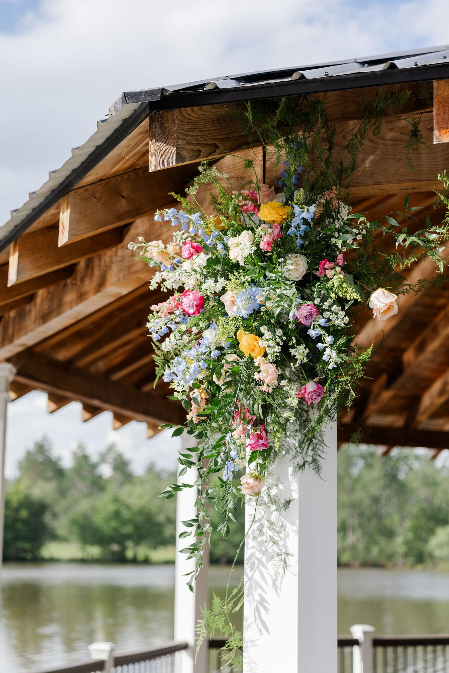 Colorful floral arrangement with blue, pink, and yellow blooms adorning a white wooden wedding ceremony pergola.