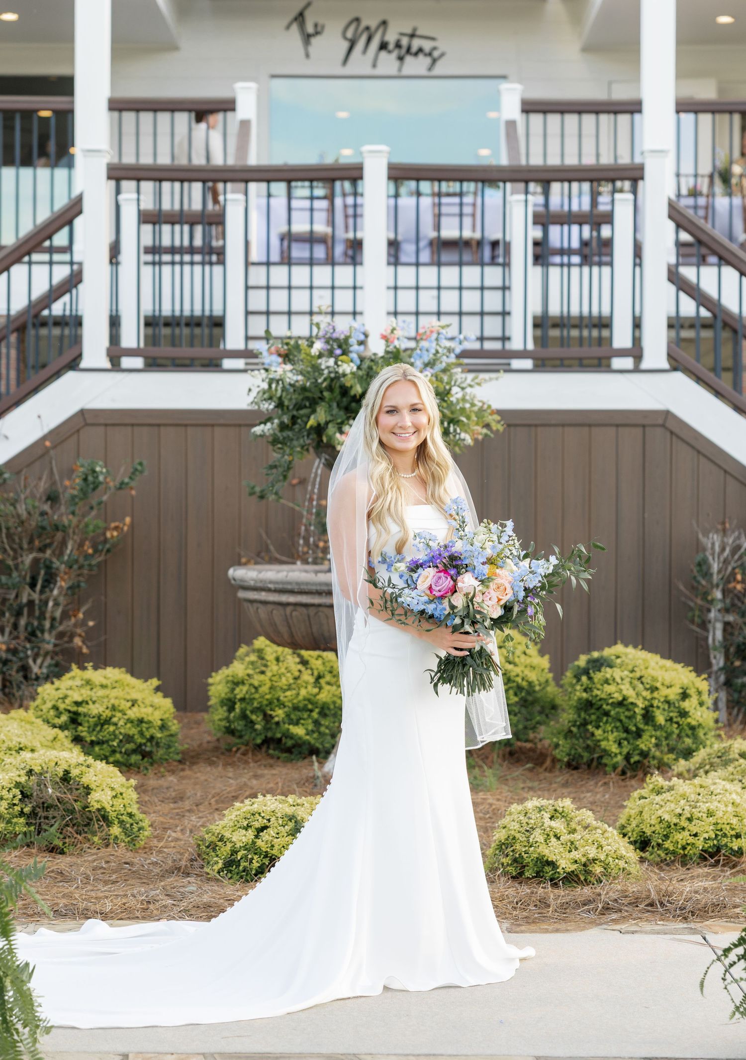 Bride in white gown holds colorful bouquet outside wedding venue with lush greenery.