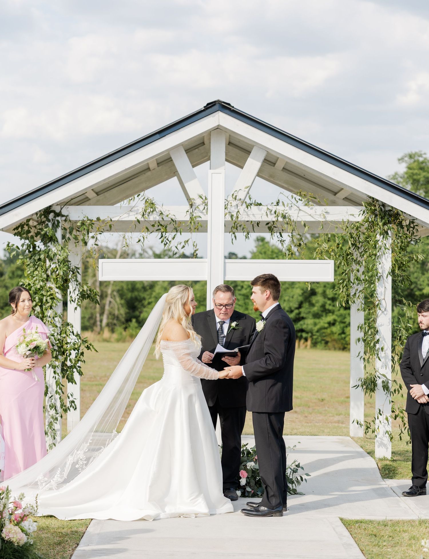 Bride and groom exchange vows at an outdoor wedding ceremony beneath a white pergola adorned with lush greenery.