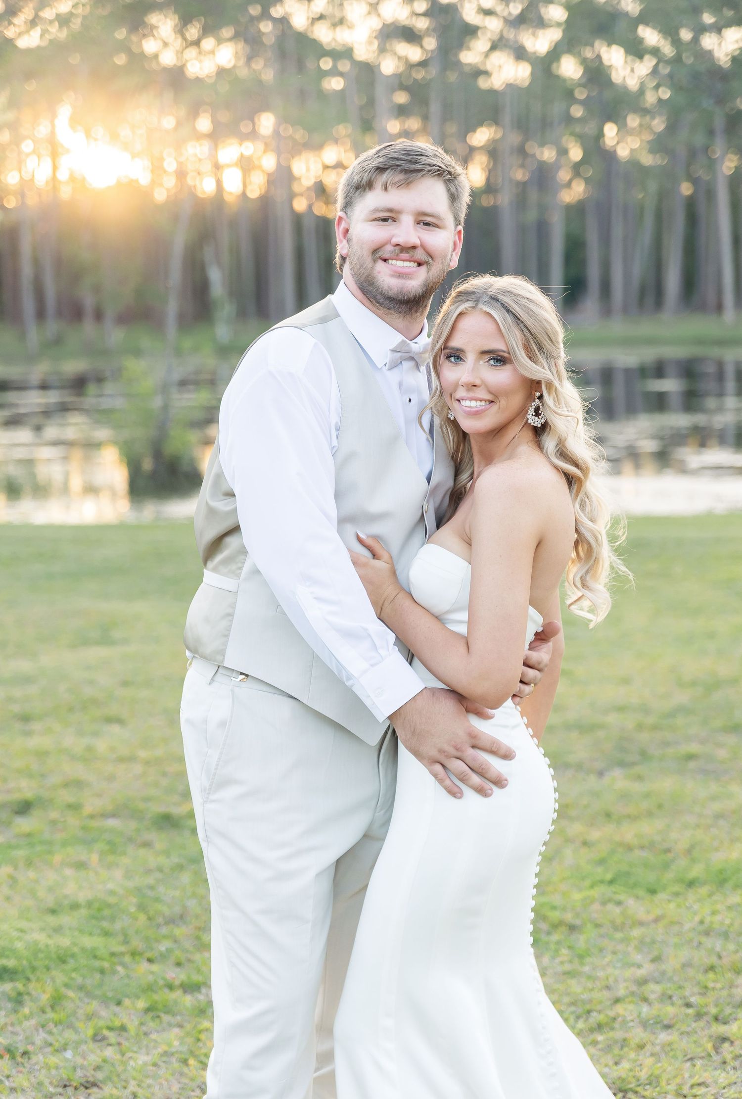 Golden hour wedding portrait featuring couple in white attire against a lakeside forest backdrop.