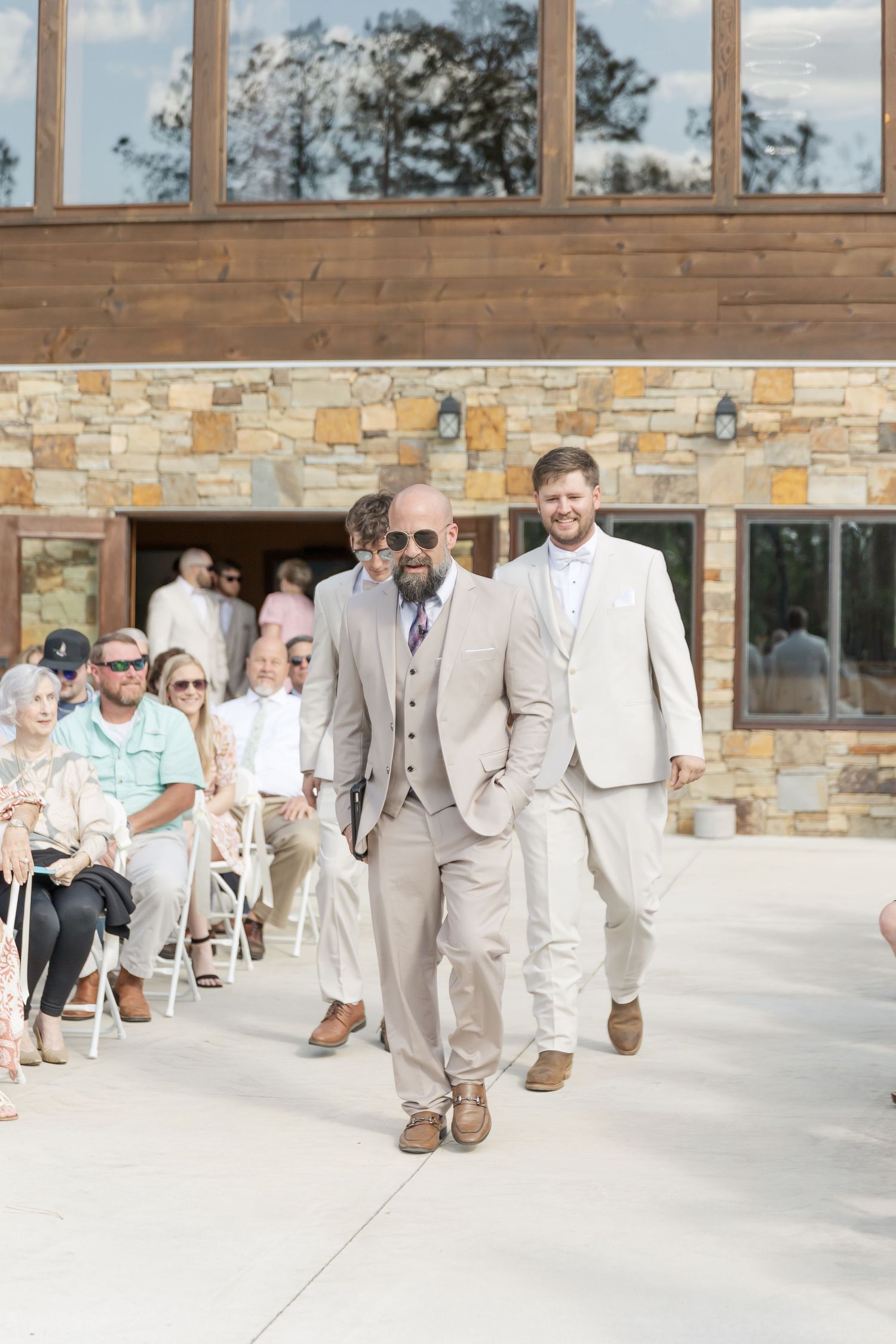 Groomsmen in beige and tan suits walk confidently down the outdoor ceremony aisle.