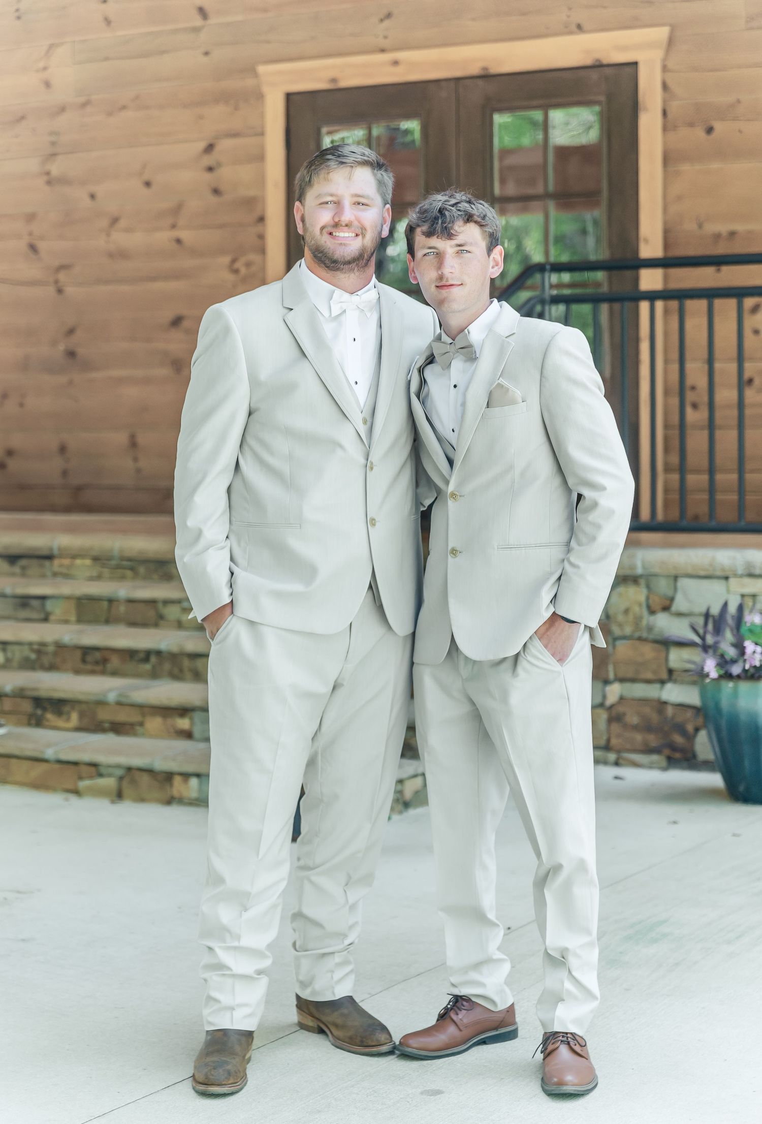 Two people in matching cream-colored suits and bow ties stand together on a patio with wooden walls behind them.