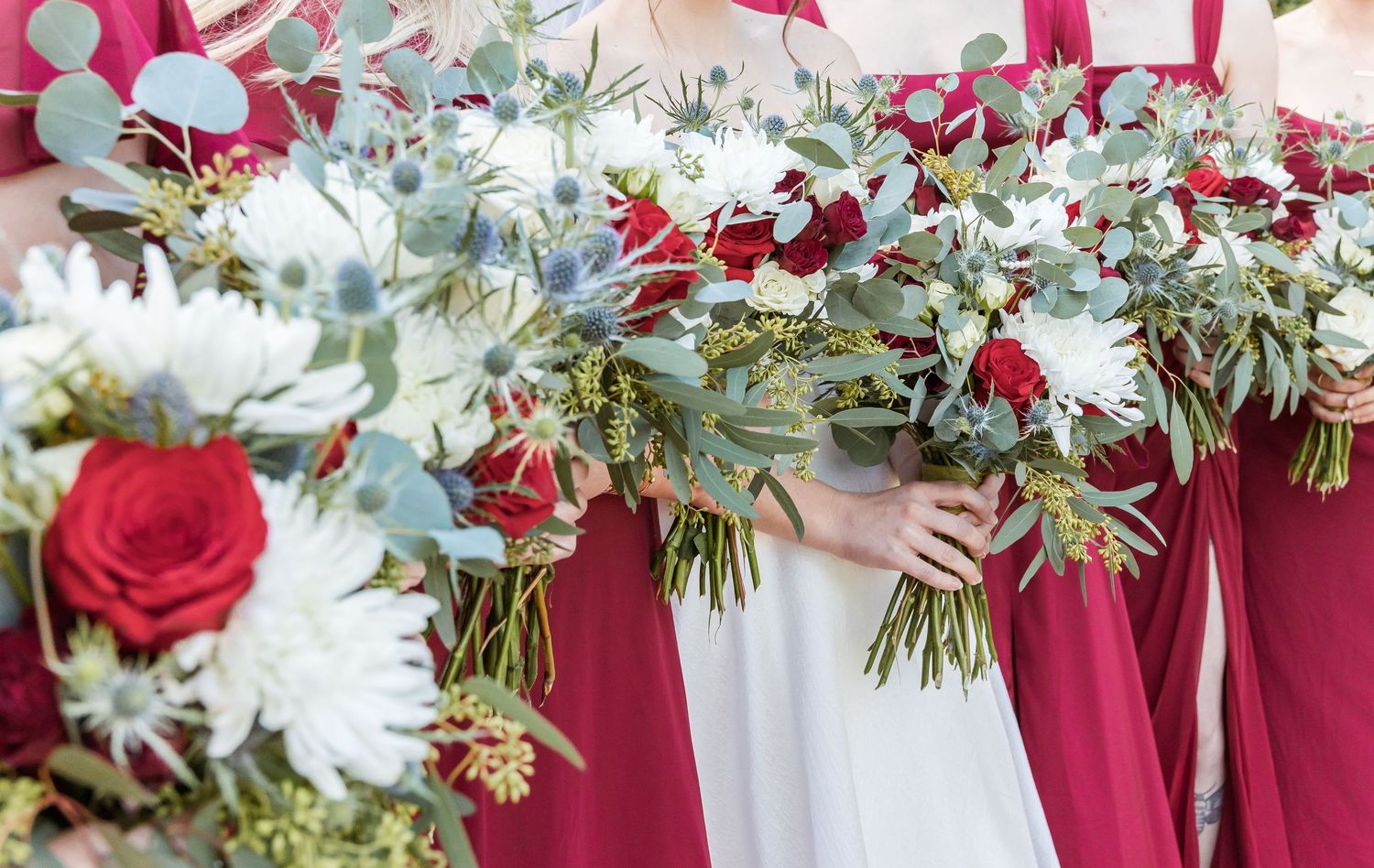 Red and white floral bouquets with greenery held against burgundy bridesmaid dresses.