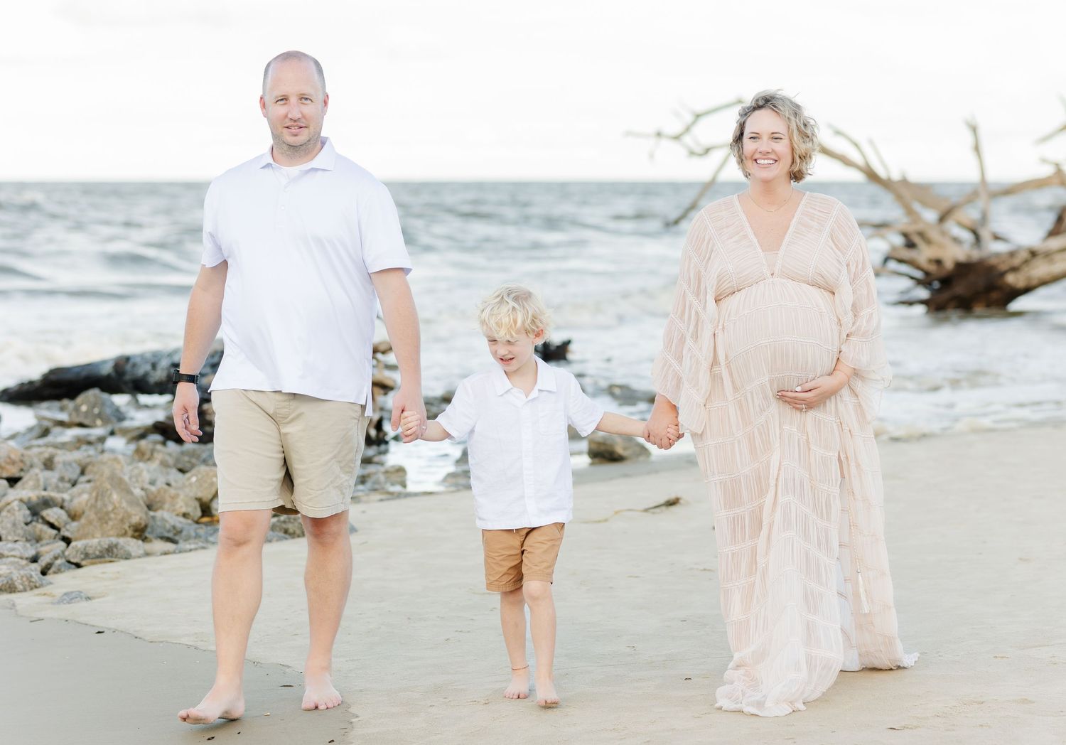 Family stands together on a rocky beach at sunset with a pregnant mother in a flowing dress and young child in white.
