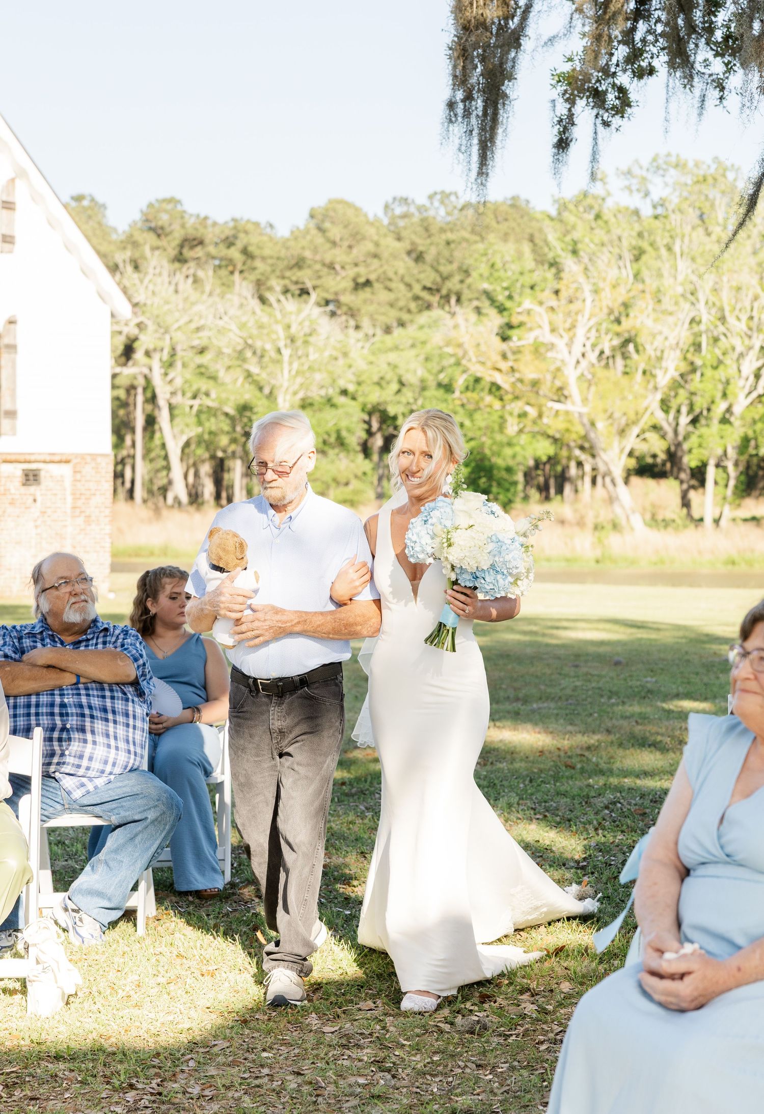 Bride in white gown holding blue bouquet walks down aisle with escort at sunny outdoor wedding ceremony.