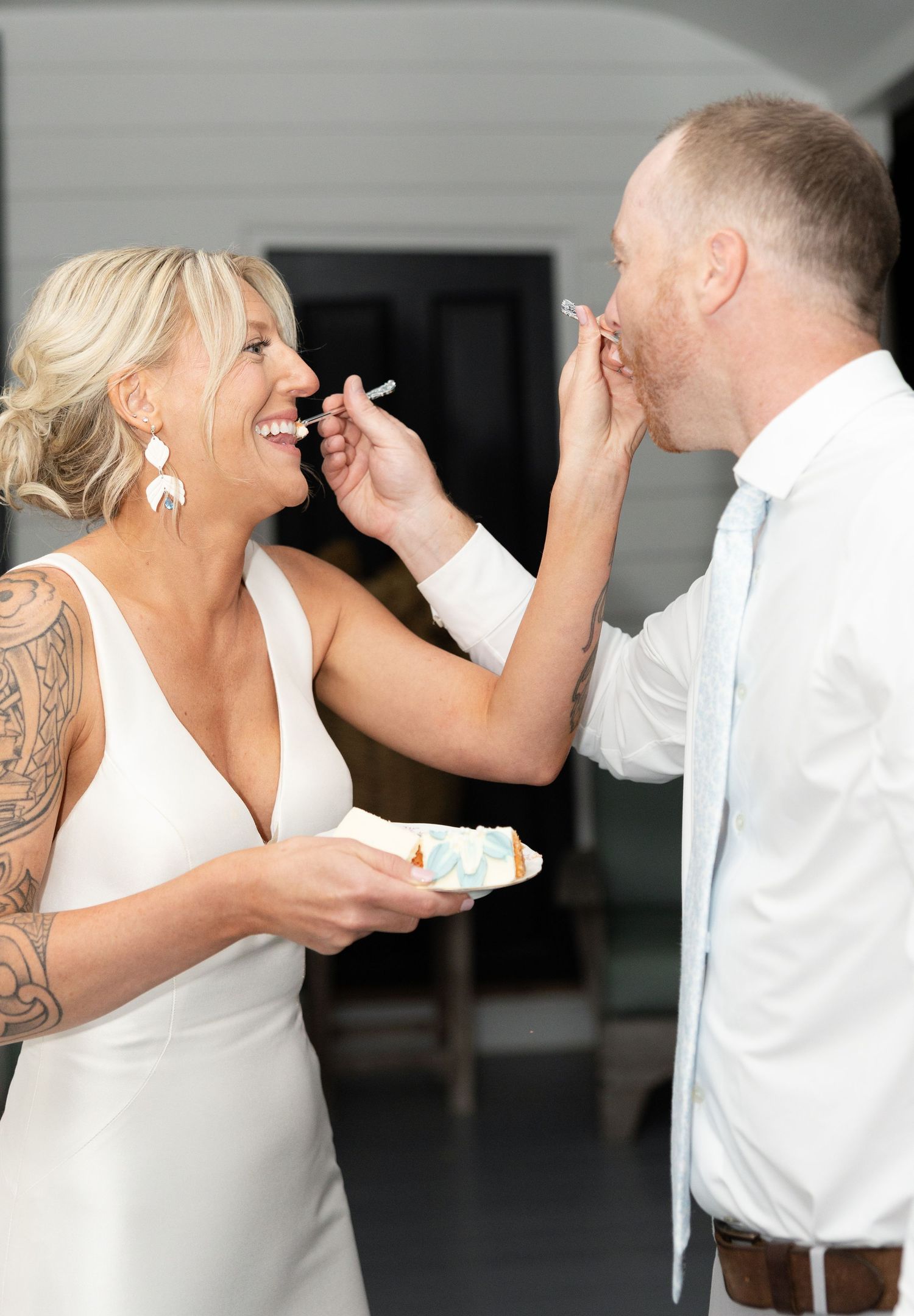 Bride with sleeve tattoo and groom playfully feeding each other wedding cake at reception.
