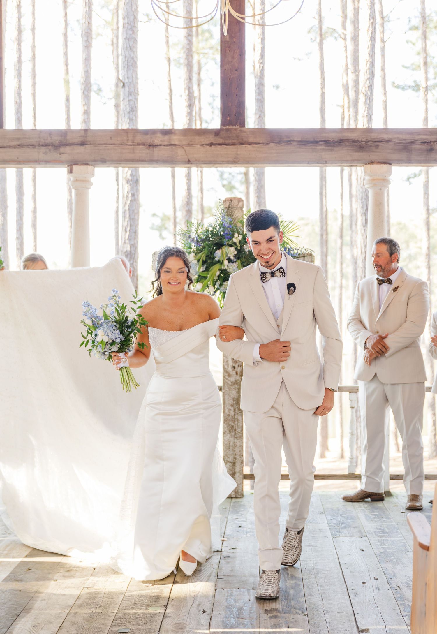 Bride in white gown and groom in cream suit walk joyfully down the aisle at a rustic wedding venue.