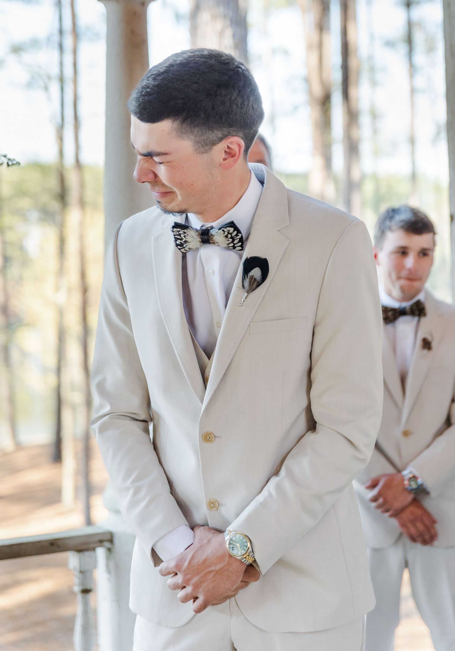 Emotional groom in cream suit with bow tie clasps hands during wedding ceremony in pine forest.
