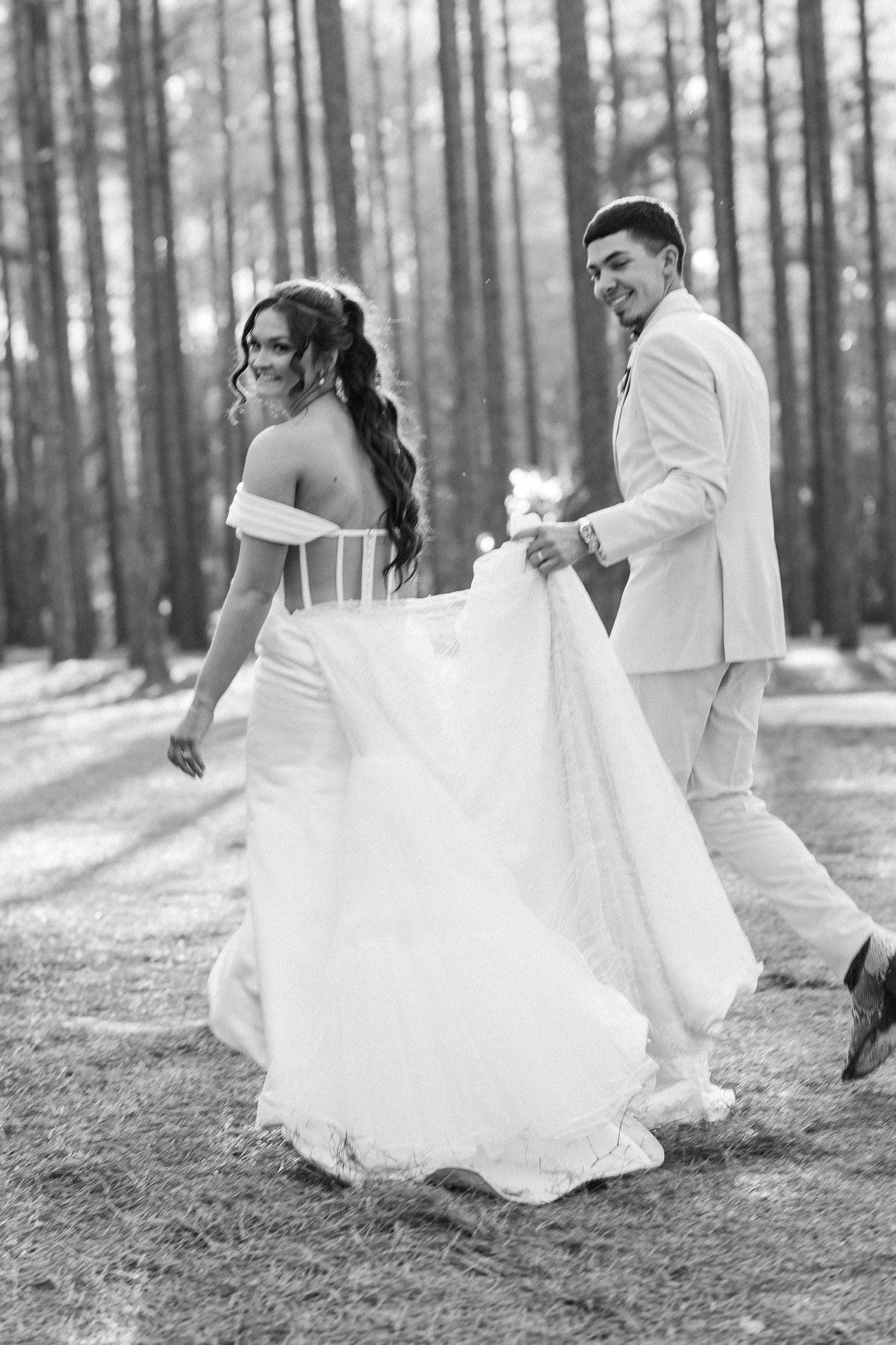 Black and white photo of smiling bride in off-shoulder gown walking with groom through pine forest.