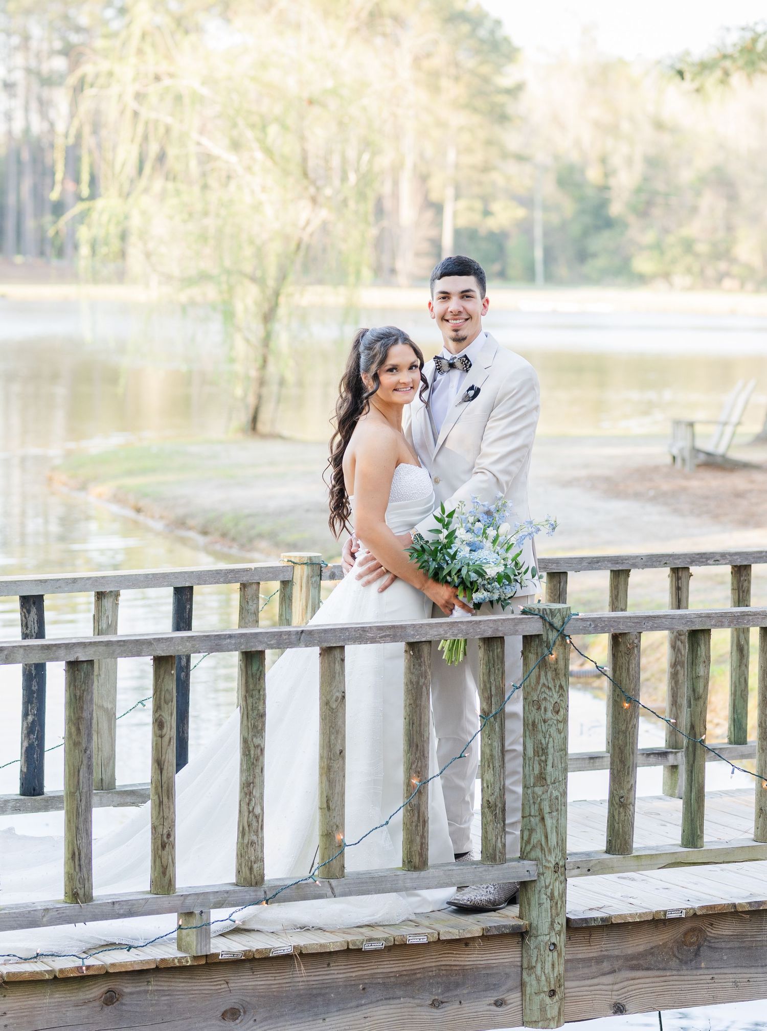 Smiling bride and groom posing on wooden dock by lake, groom in beige suit holding blue bouquet.