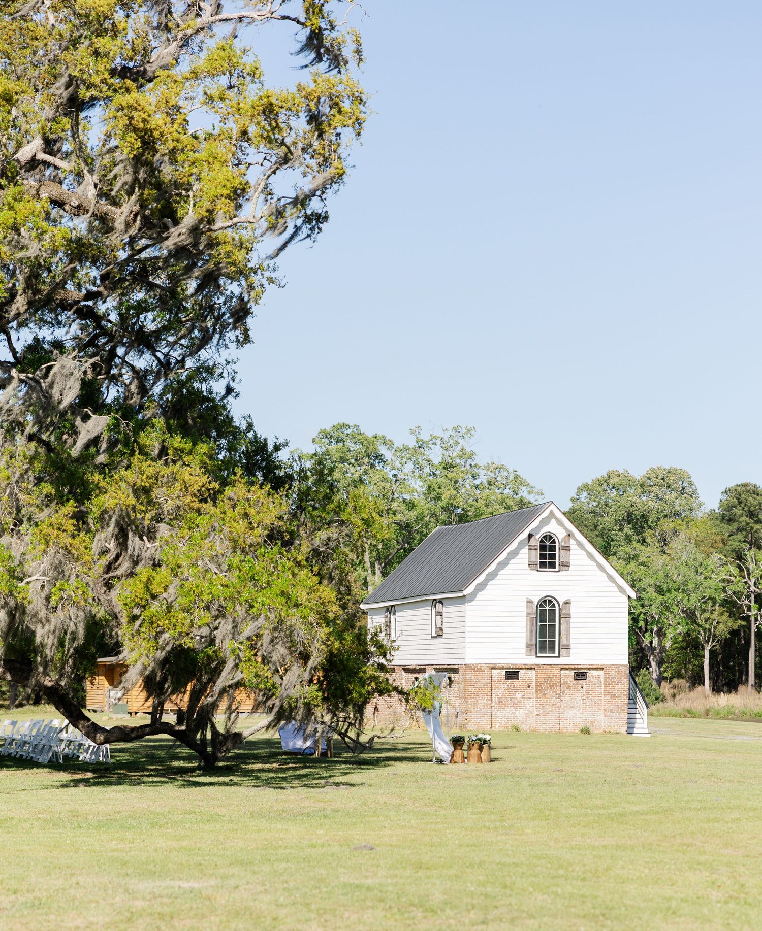 White chapel with brick foundation surrounded by Spanish moss trees on sunny wedding venue grounds.