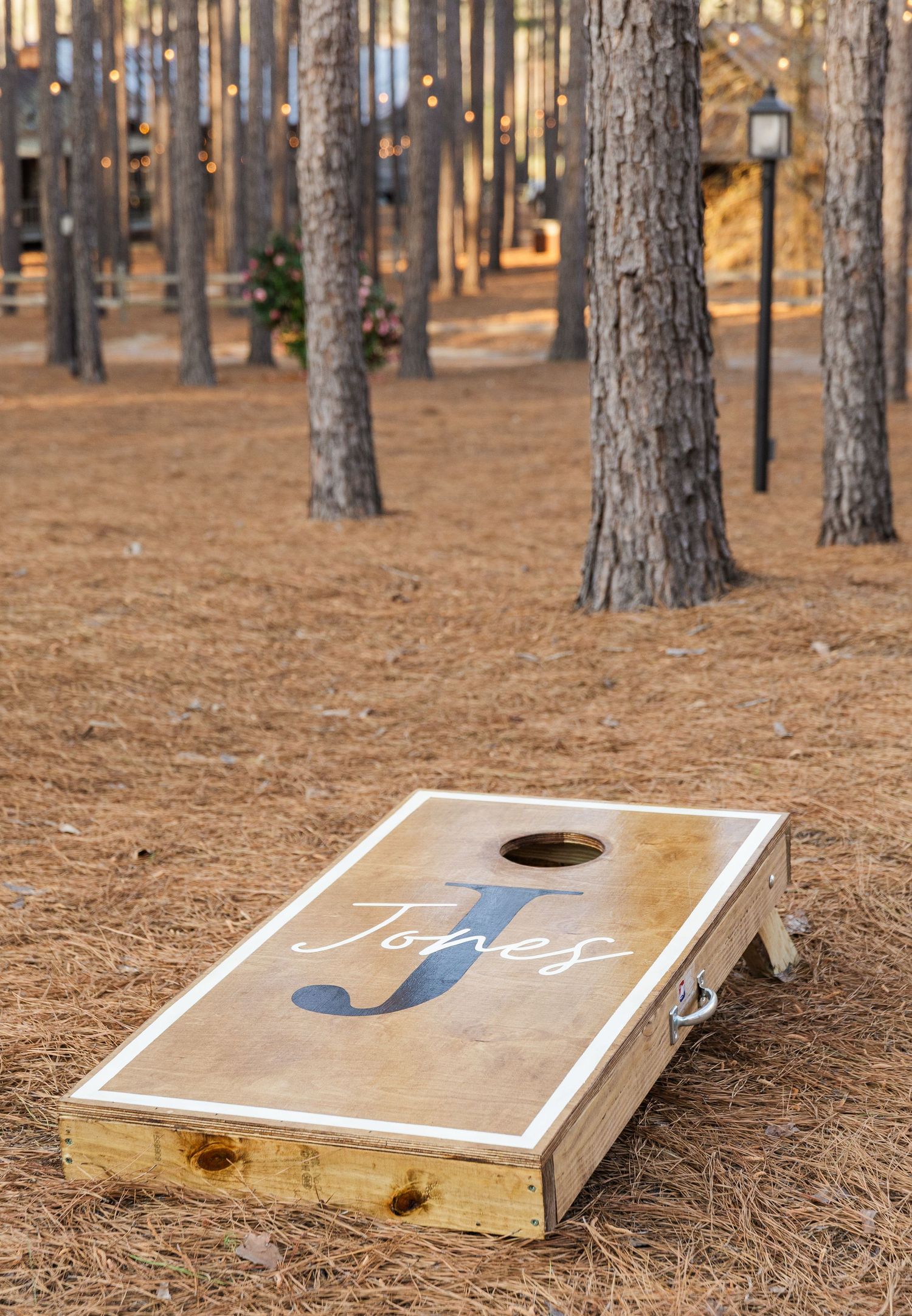 Custom wooden cornhole board with Jones monogram displayed among pine trees with string lights at an outdoor wedding.