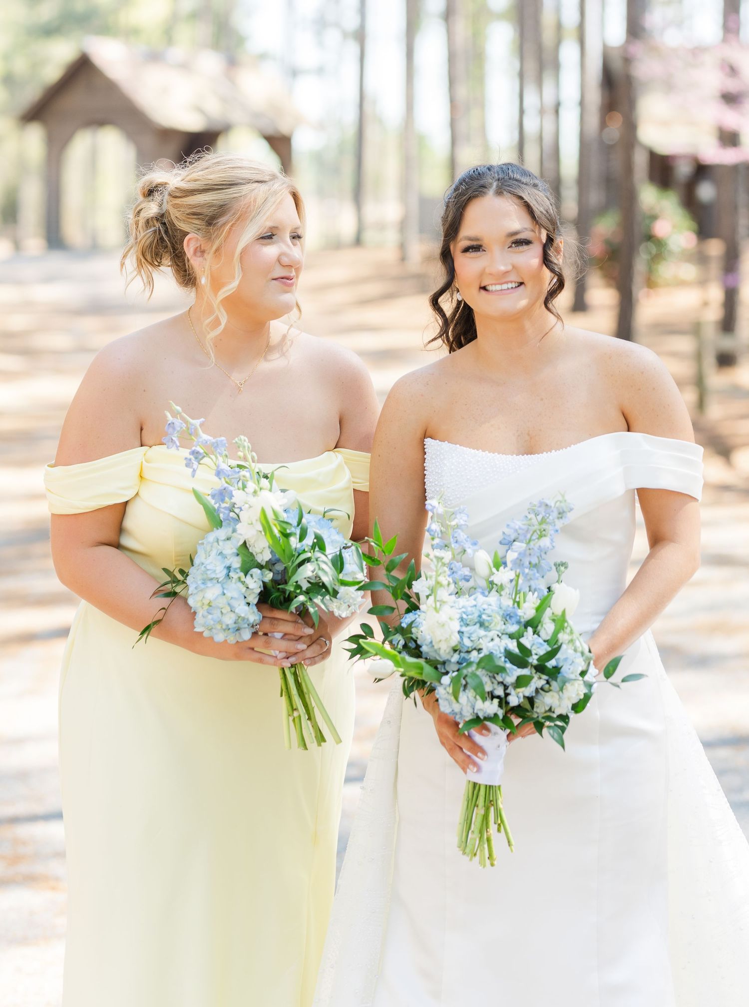 Bride and bridesmaid in yellow gown hold matching blue floral bouquets outdoors at wedding.
