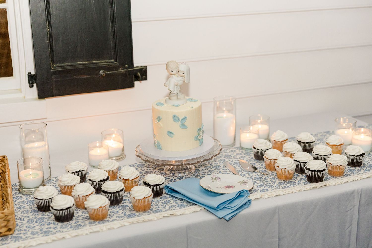 Wedding dessert table with small white cake topped with mouse figurine, cupcakes, and glowing candles.
