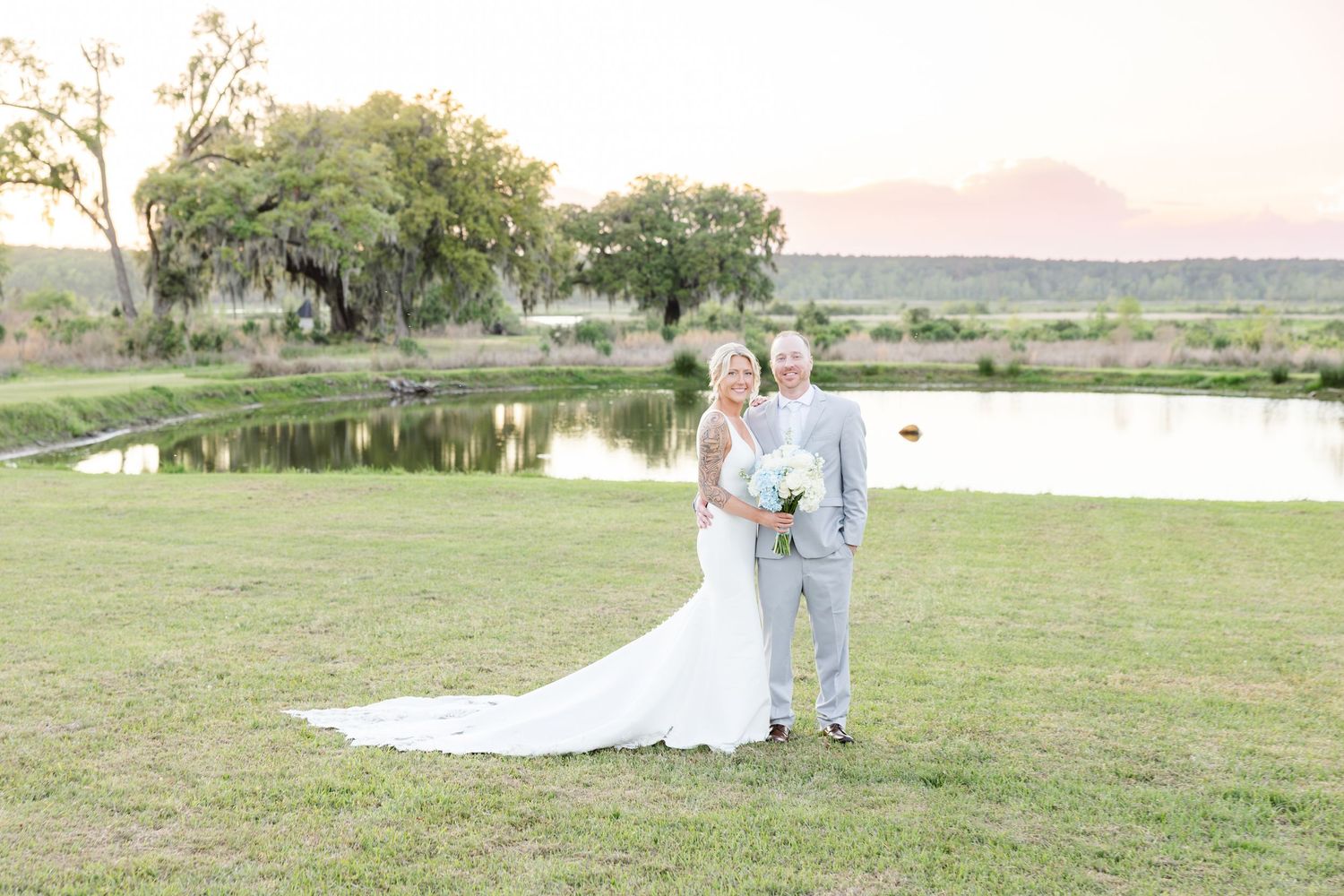 Bride and groom posing on green lawn at sunset with scenic pond and mountains in background.
