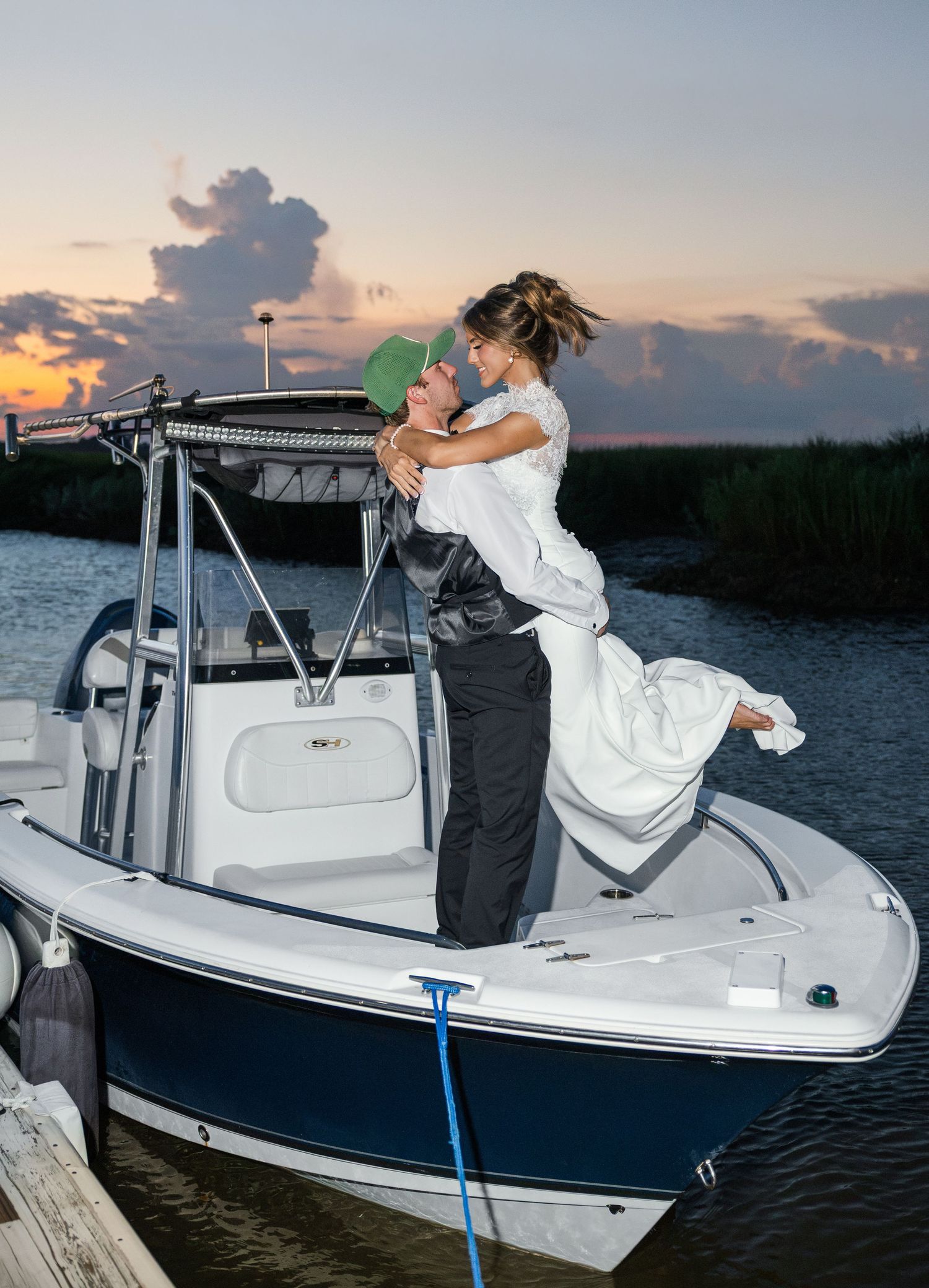 Couple embracing on a white motorboat at sunset, woman in wedding dress lifted by groom near a dock.
