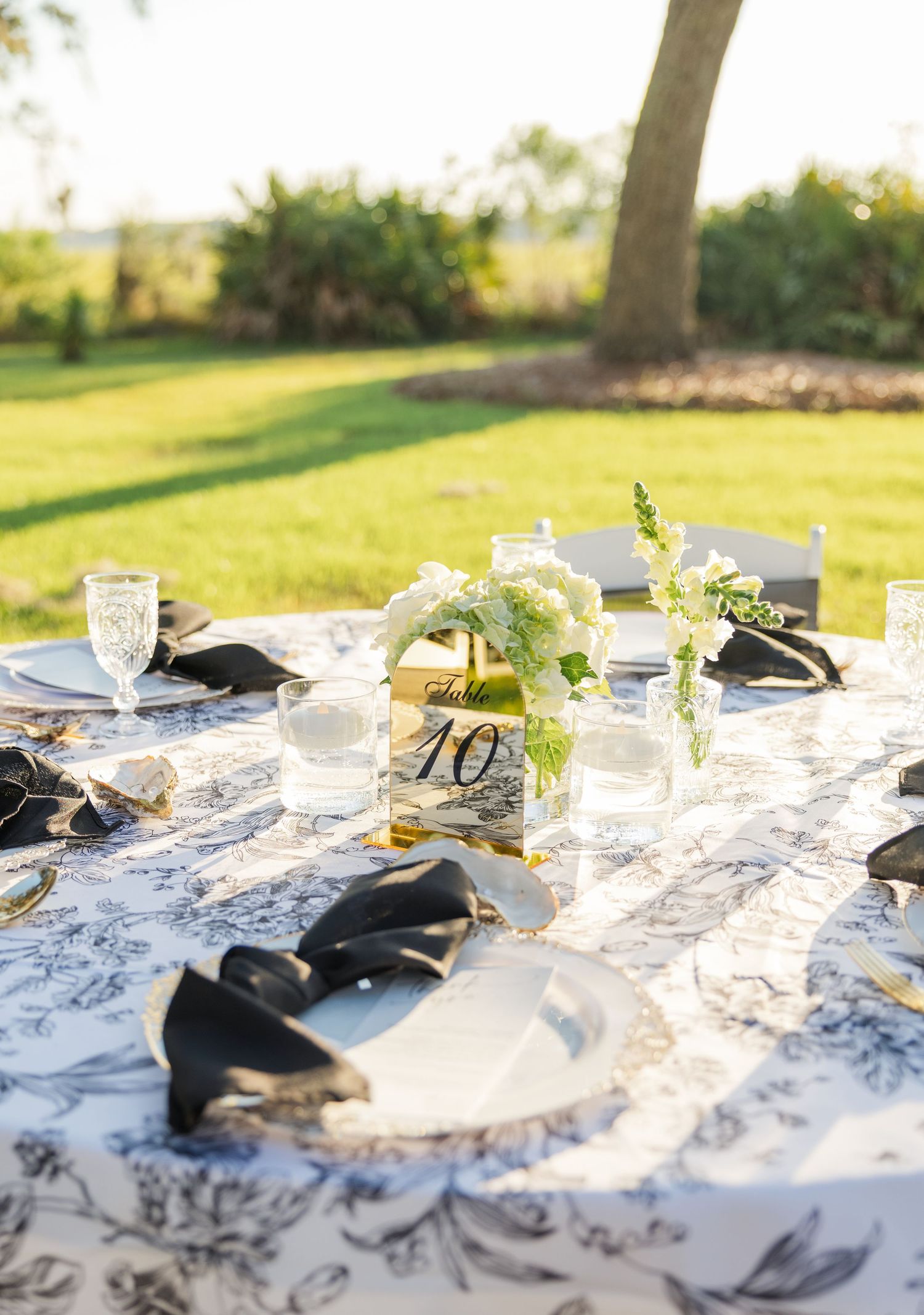 Outdoor wedding table with black floral toile linen, gold chargers, and white floral centerpieces.