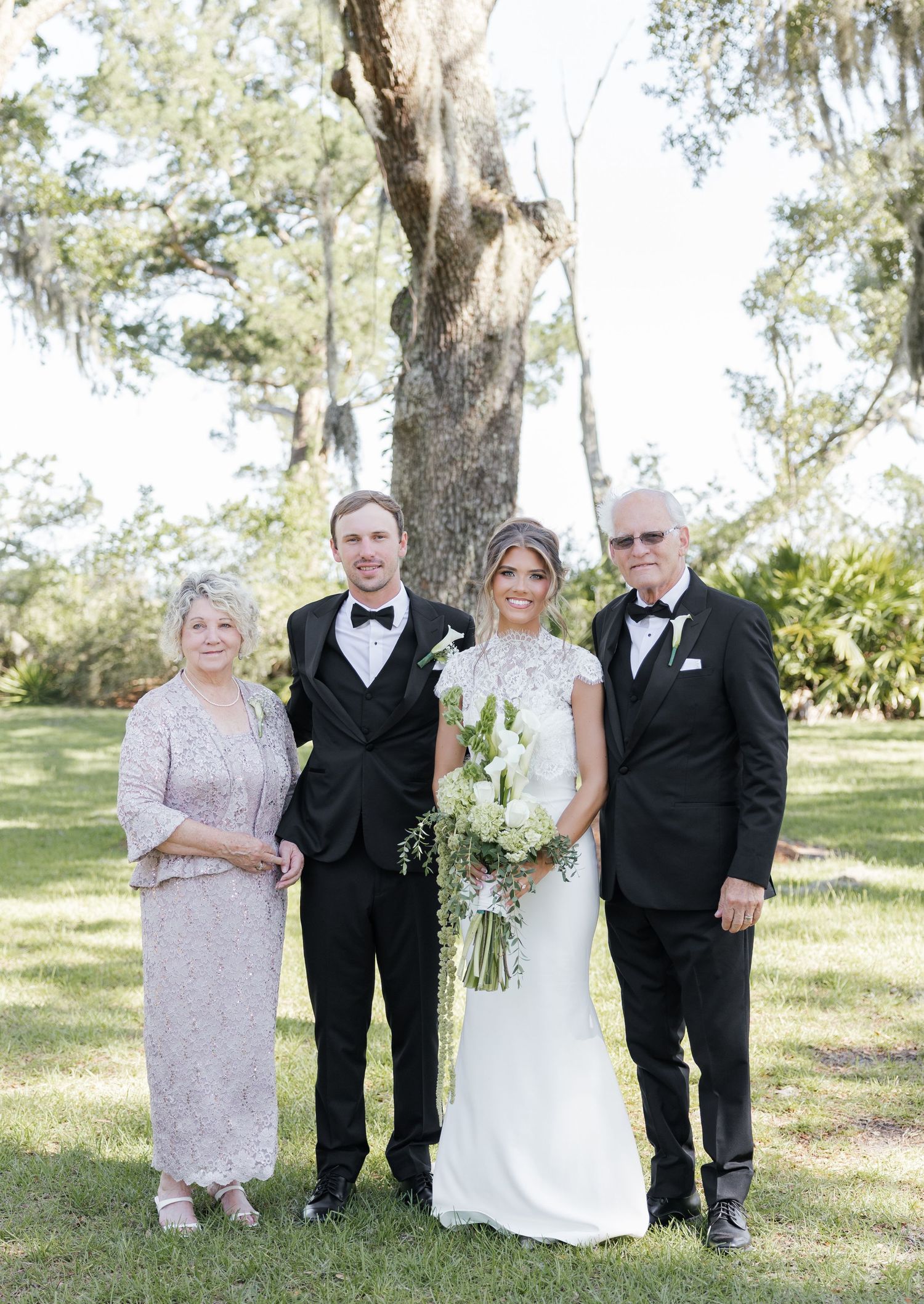 Wedding party of four posing outdoors under oak trees, bride holding white bouquet in elegant gown with groom and parents.