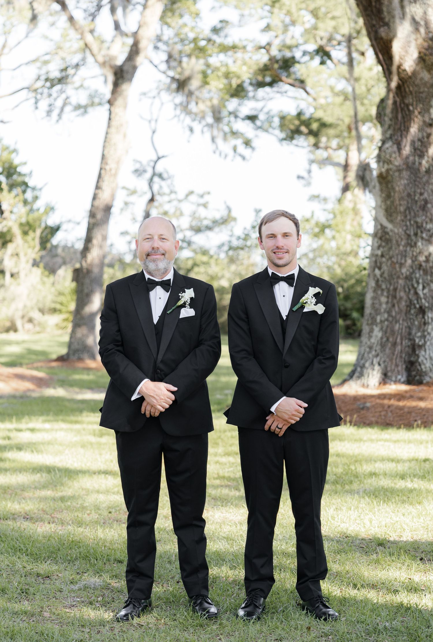 Two men in black tuxedos with white boutonnieres stand smiling outdoors on green grass beneath large oak trees.