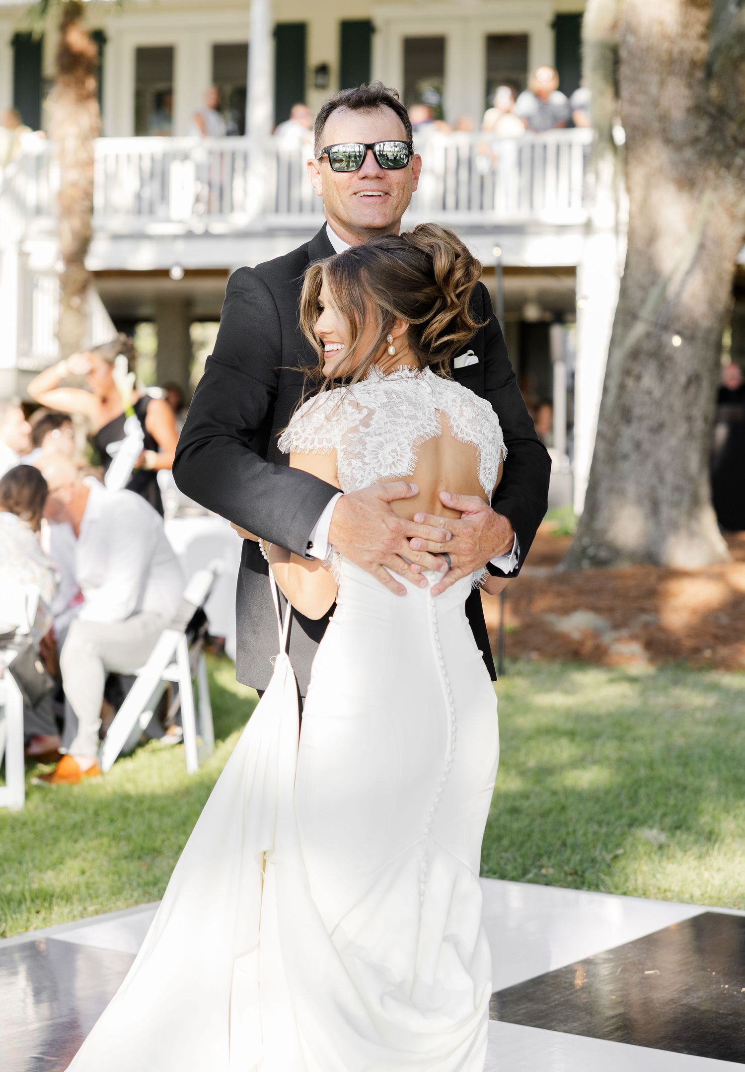 Groom in sunglasses and black suit holds bride in lace wedding dress during outdoor first dance ceremony.
