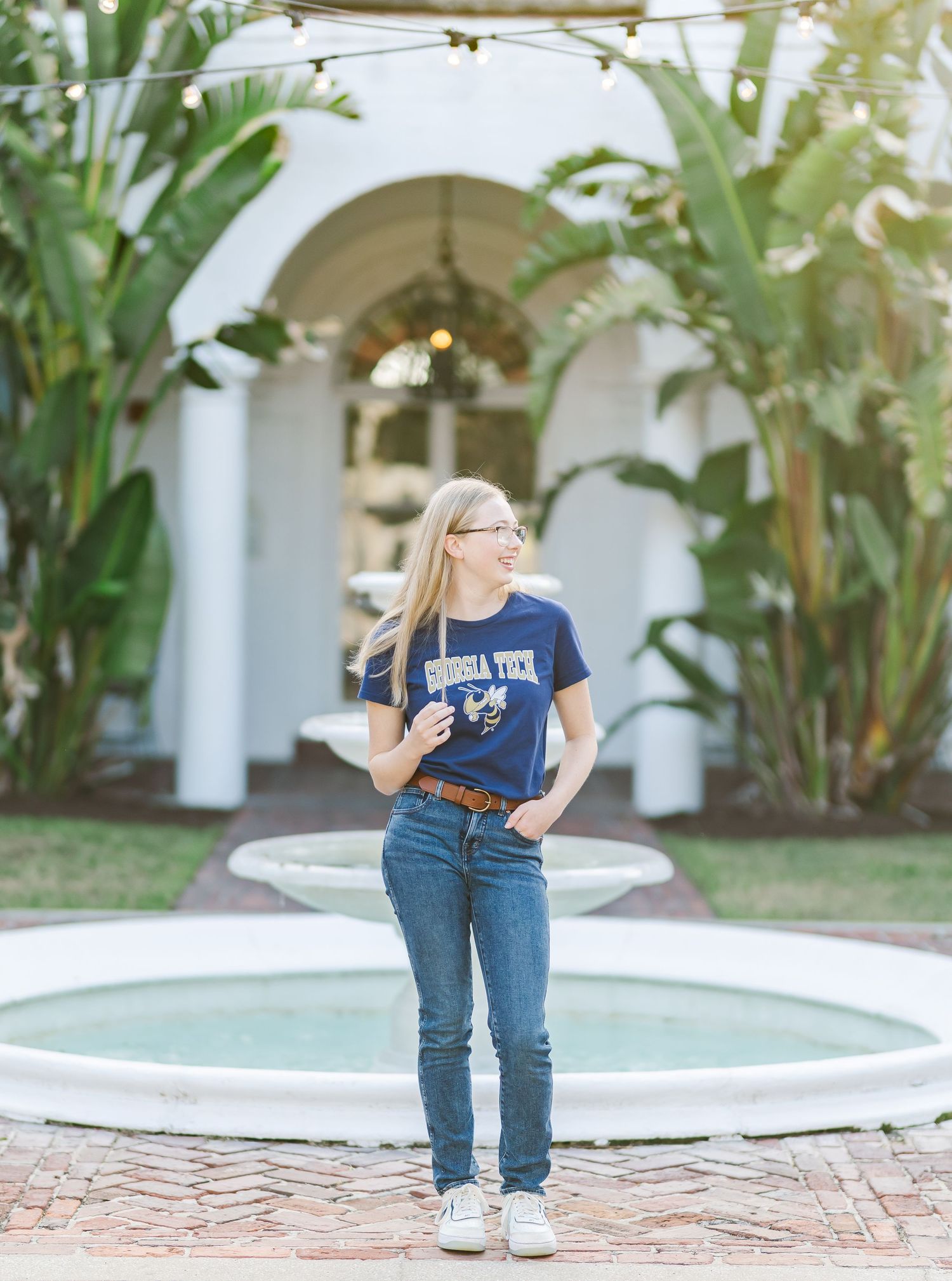 Person wearing navy top and jeans poses casually near tropical plants and white archways in outdoor courtyard setting.