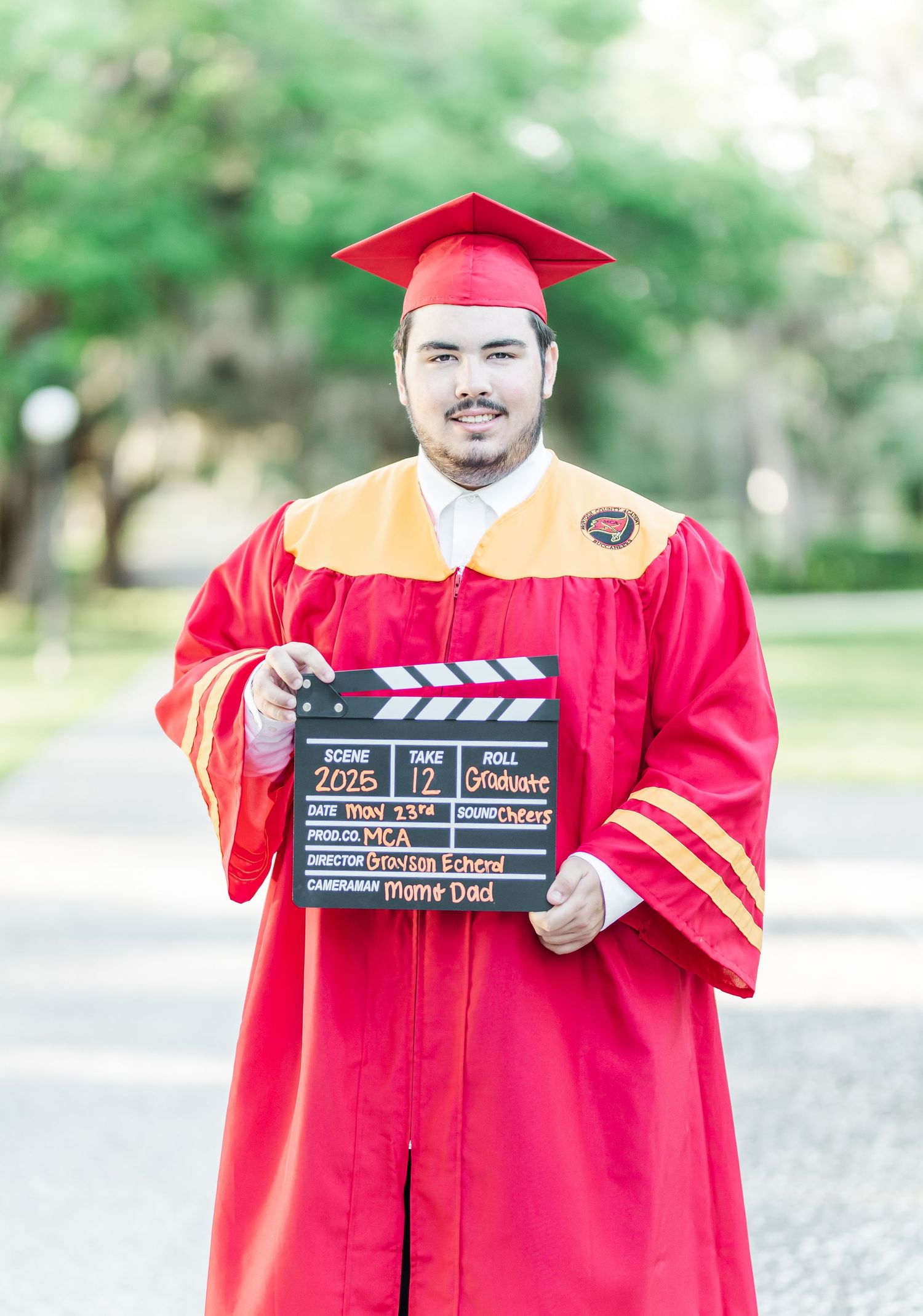 Graduate in red cap and gown holds a black film clapperboard during outdoor photo session.