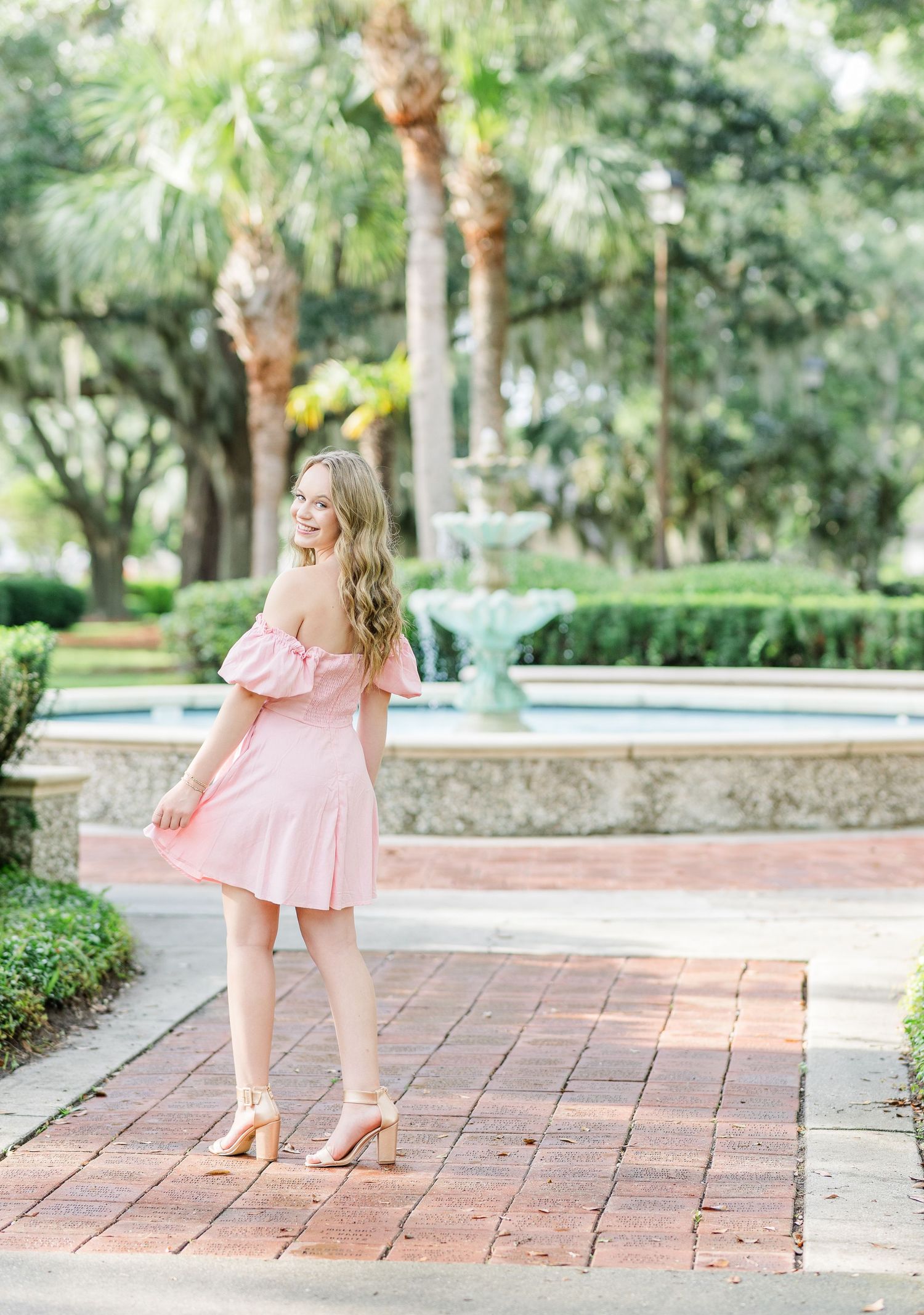 A person in a pink dress and heels stands on brick pavers near a decorative fountain.