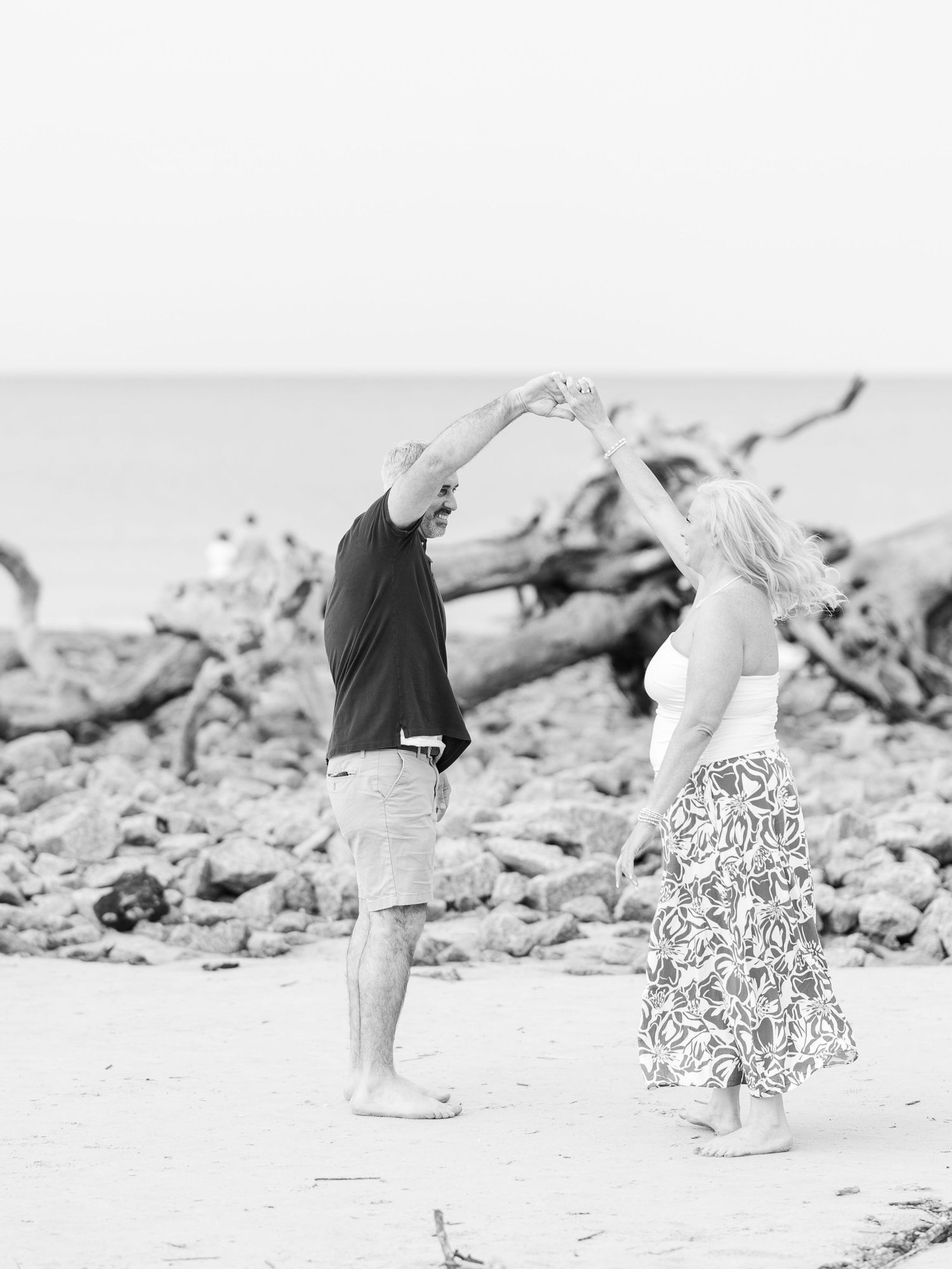 A couple dances together barefoot on a beach with driftwood in black and white.