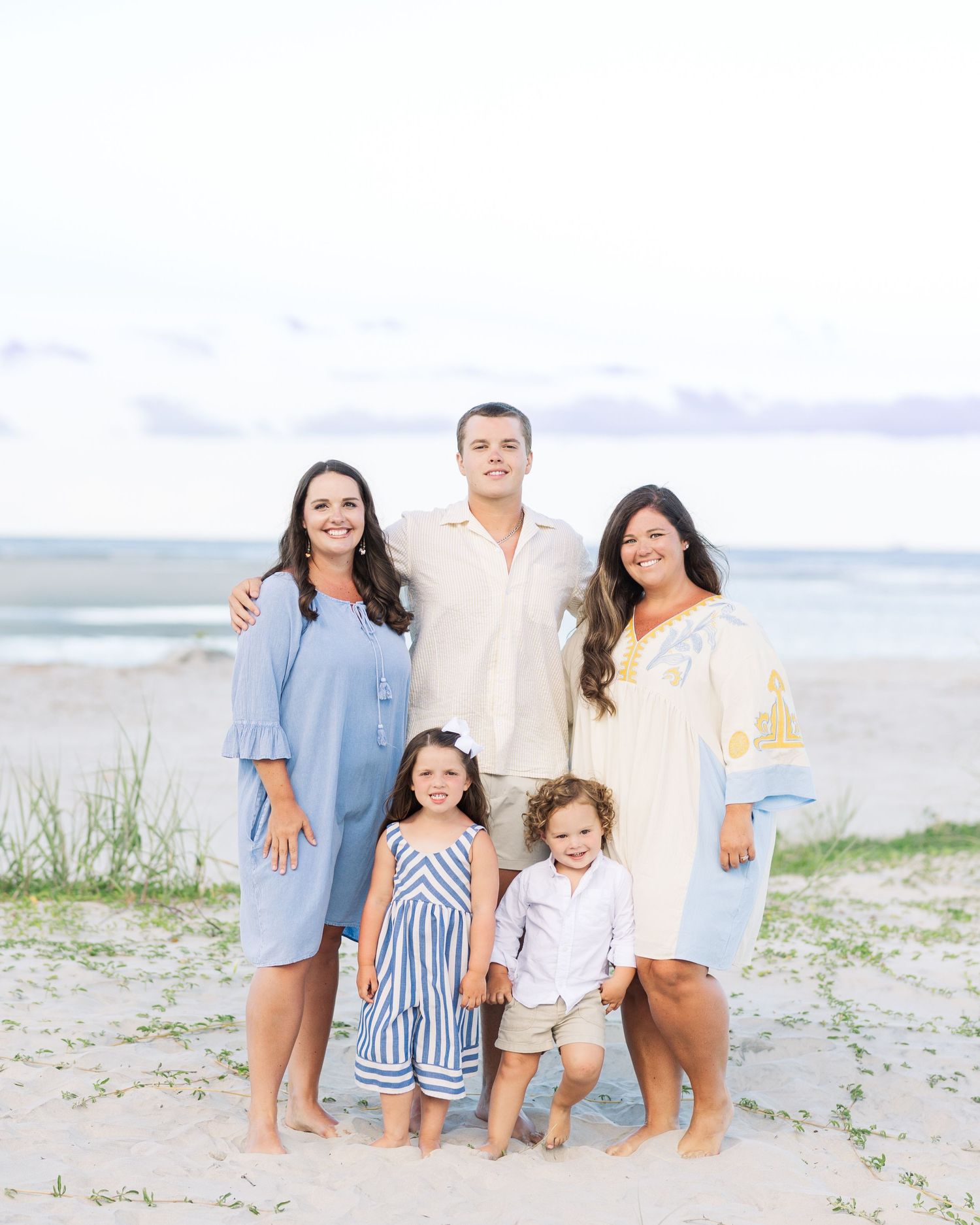 A happy family wearing coordinated blue and white outfits poses together on a sandy beach at sunset.
