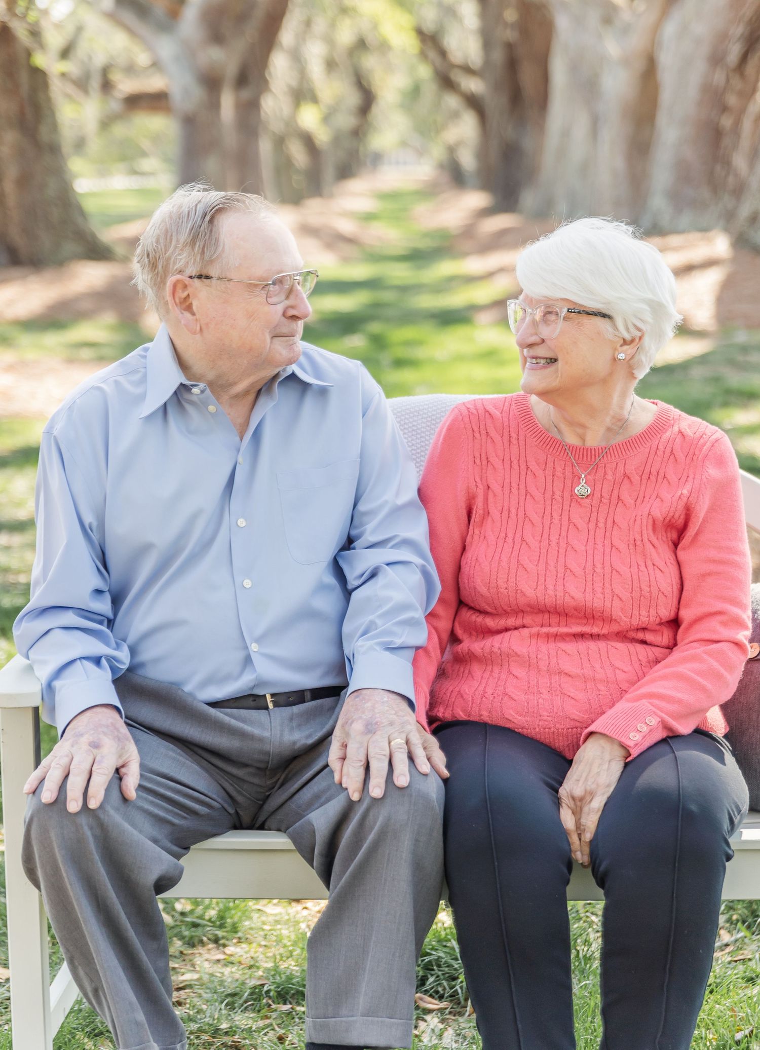 An elderly couple sits together on a bench, sharing a joyful moment in a park setting.